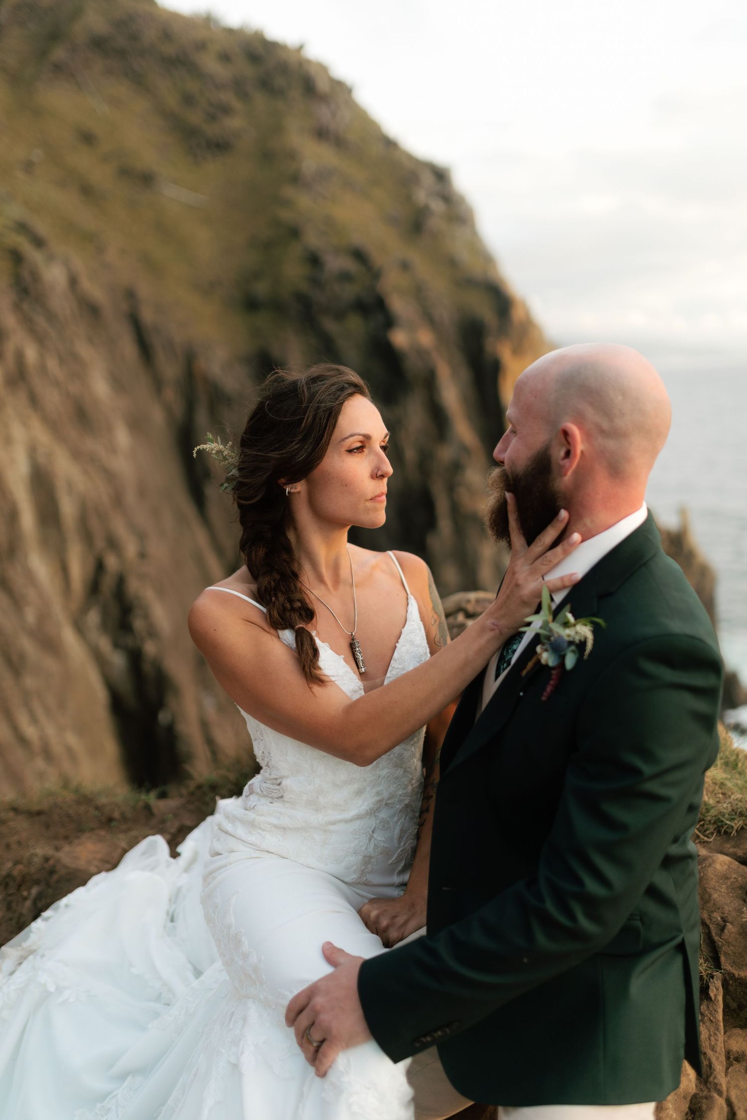 Bride tenderly touching grooms face while seated on rocky coastal cliffs.