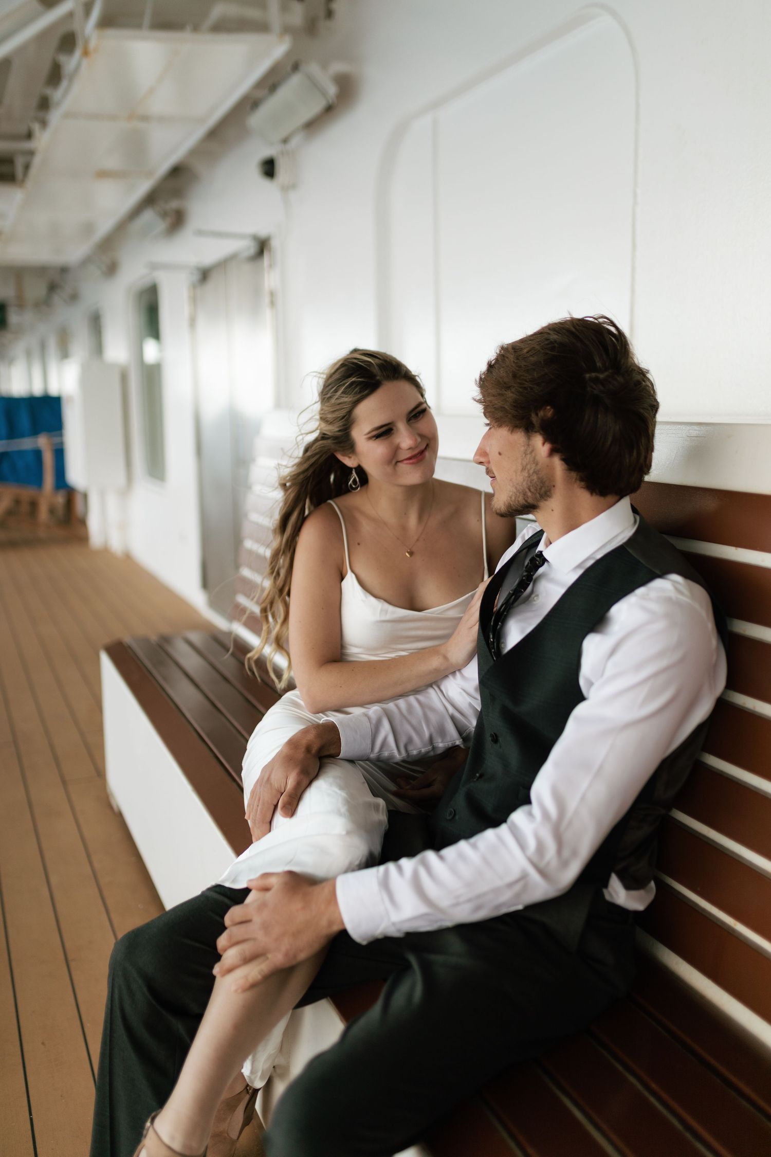 Smiling bride and groom sit closely together on a wooden bench on a cruise ship deck.