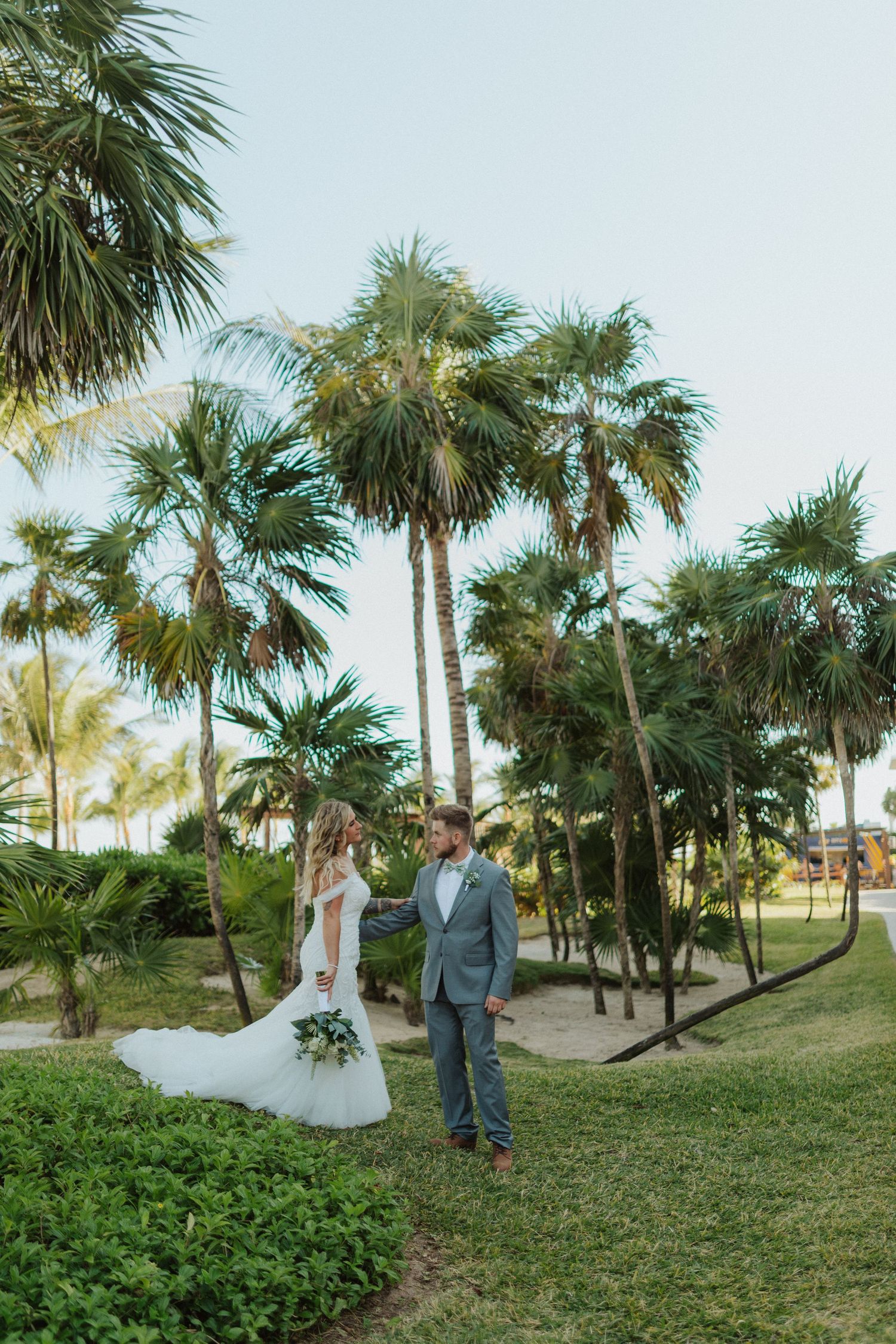 Bride and groom walking hand in hand through lush tropical garden with tall palm trees at sunset.