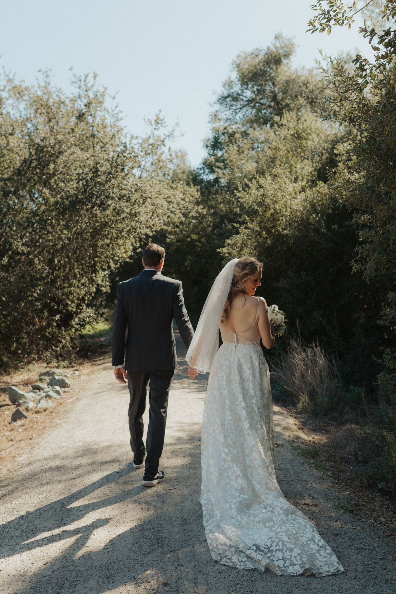 Bride and groom walk hand in hand down a sunlit tree-lined path after their ceremony.
