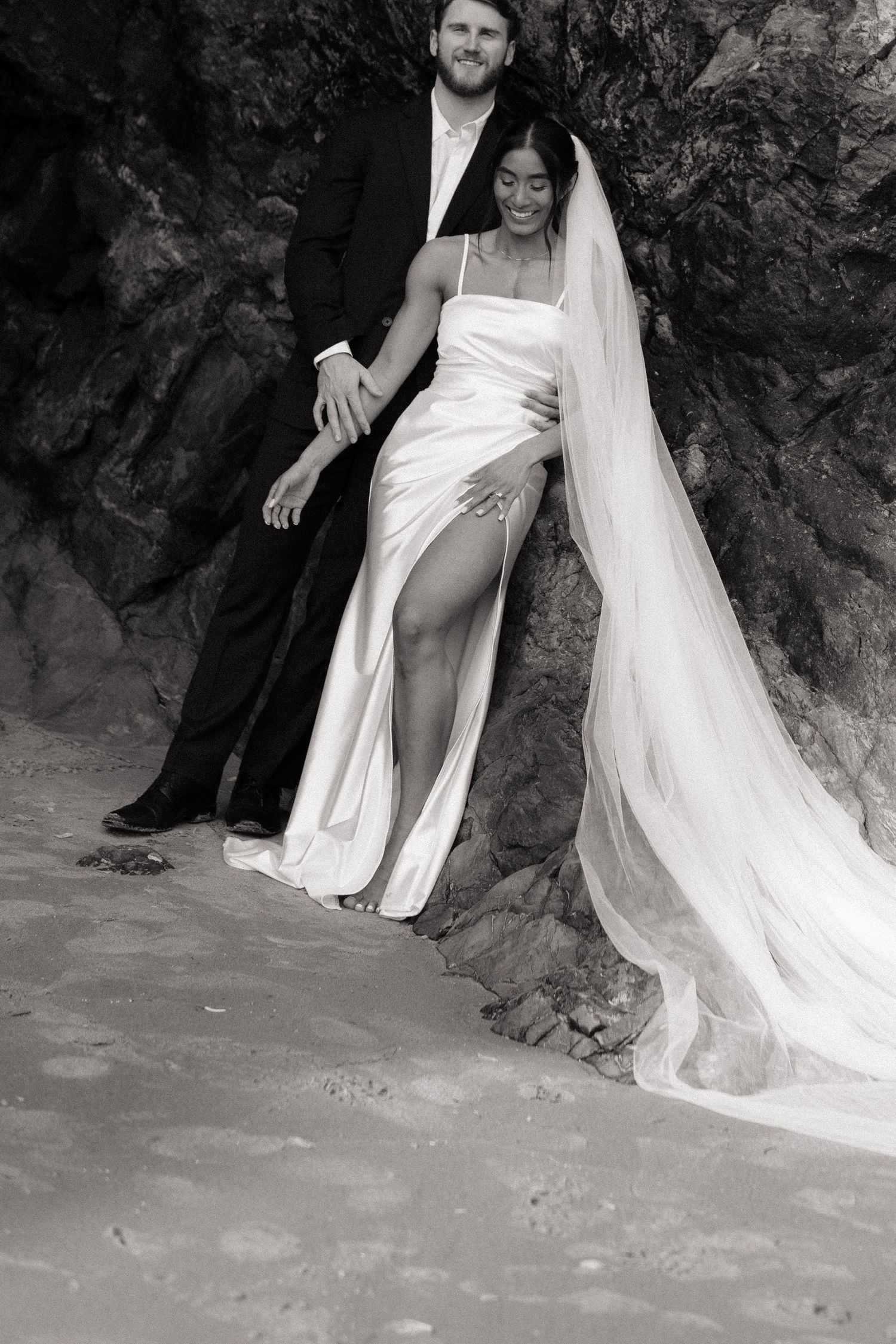Black and white portrait of bride and groom leaning against coastal rock, bride in satin dress with flowing veil.