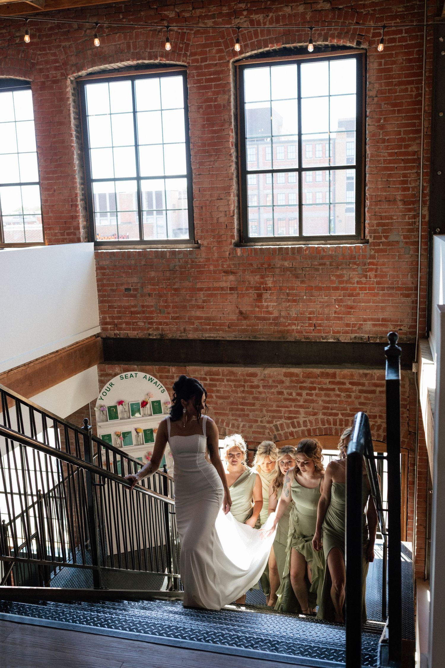Bride in white gown leads bridesmaids in sage green up iron staircase in brick industrial venue.