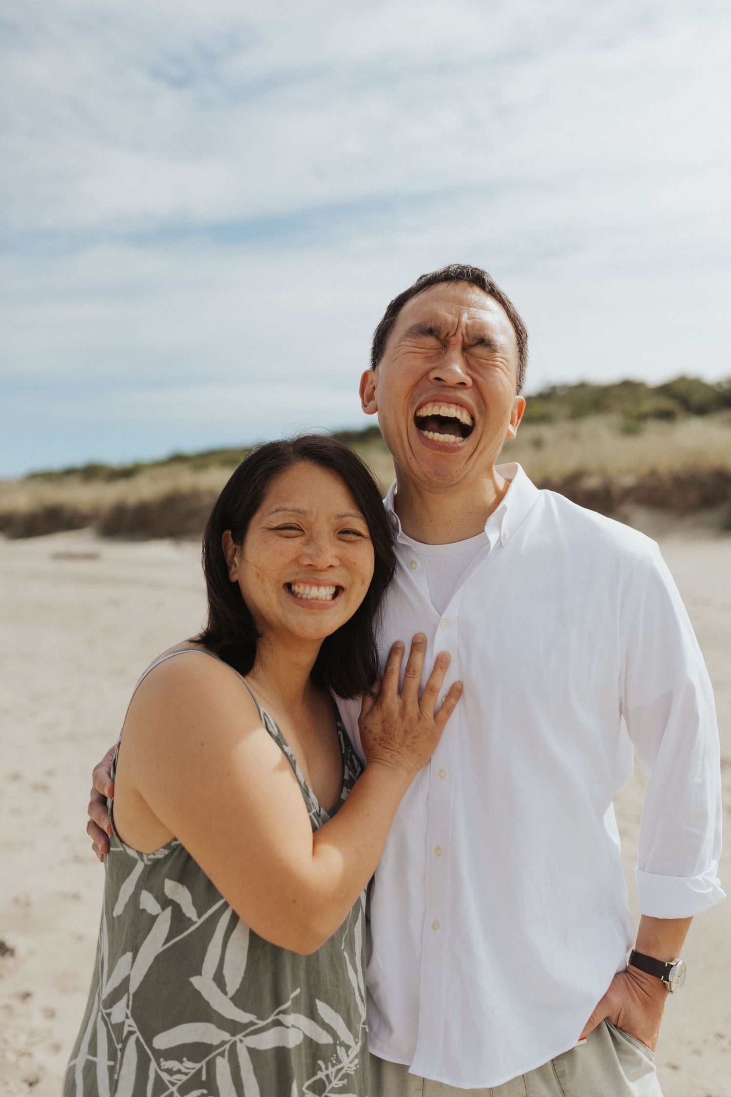 Joyful Asian couple laughing and embracing on a sandy beach under a cloudy sky.