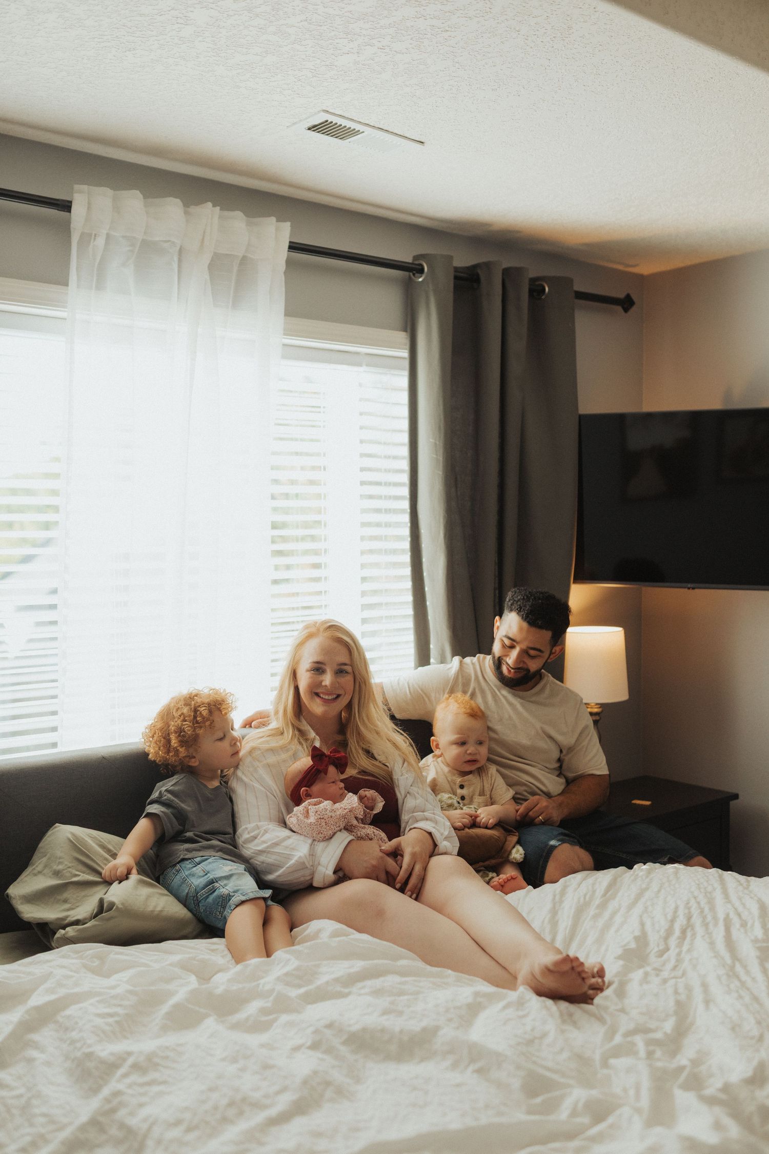 A smiling blonde mother holds a newborn surrounded by her family on a large white bed.