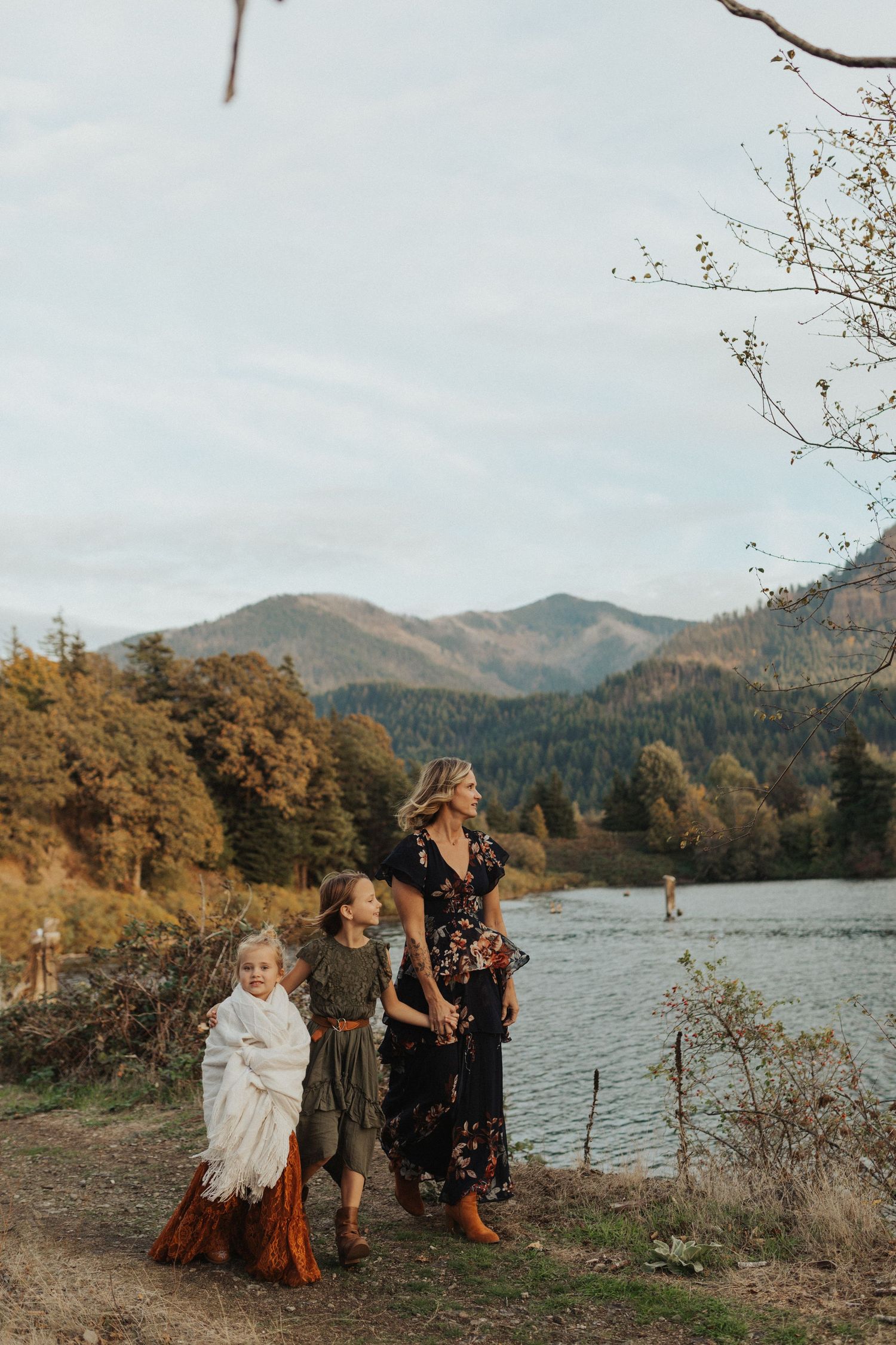 A mother and two daughters stand by a mountain lake on an autumn day, wearing bohemian dresses.