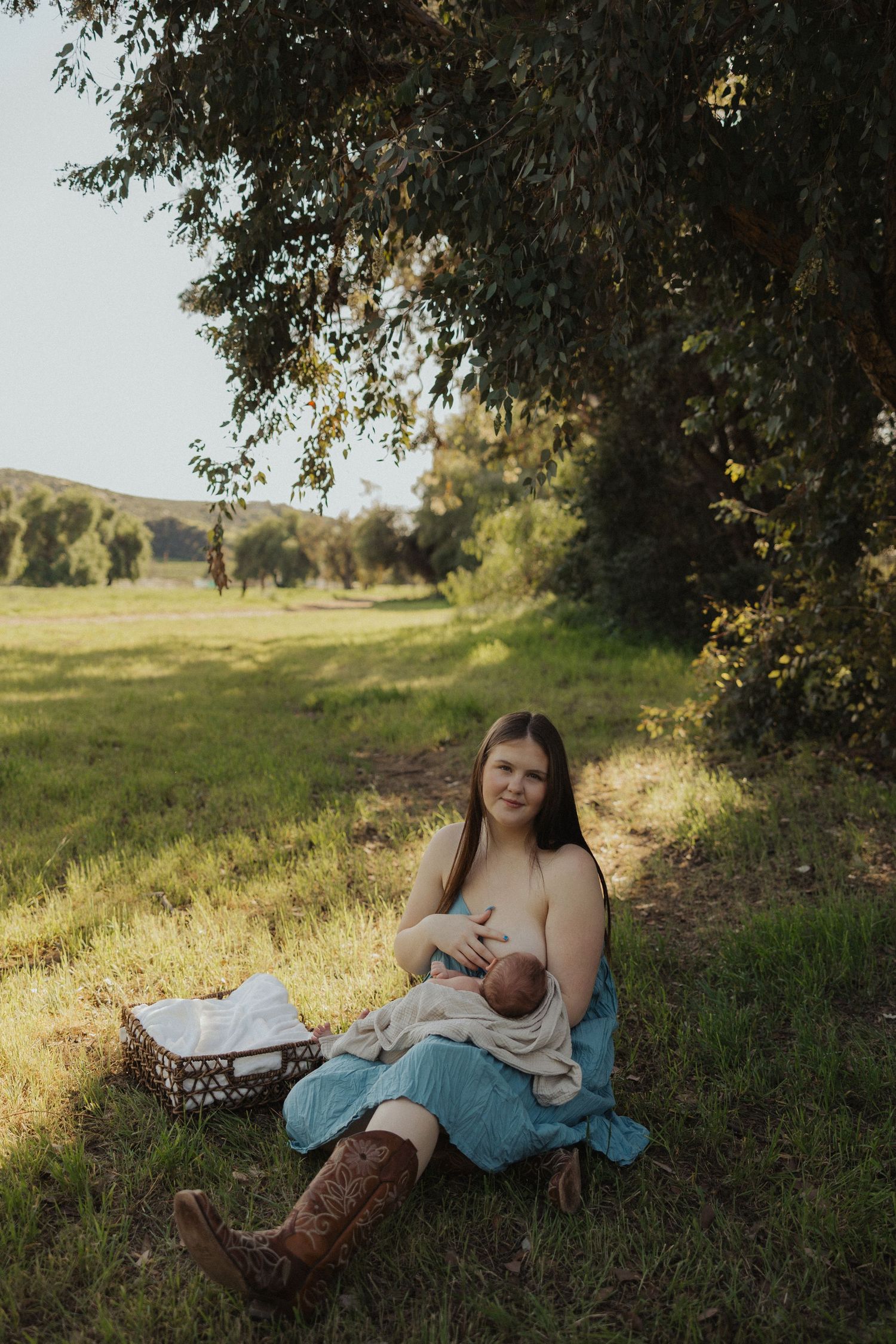 Mother in teal dress breastfeeding newborn baby while sitting on grass under trees in golden sunlight.