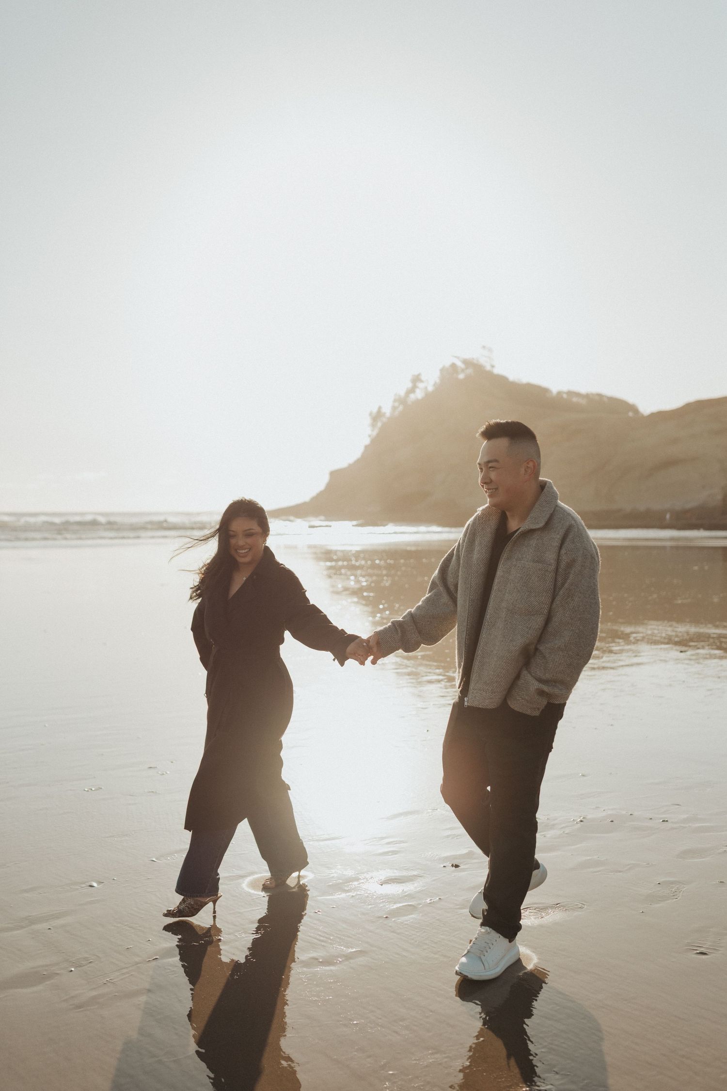 Newly engaged couple holds hands walking on reflective wet sand beach at golden hour sunset.