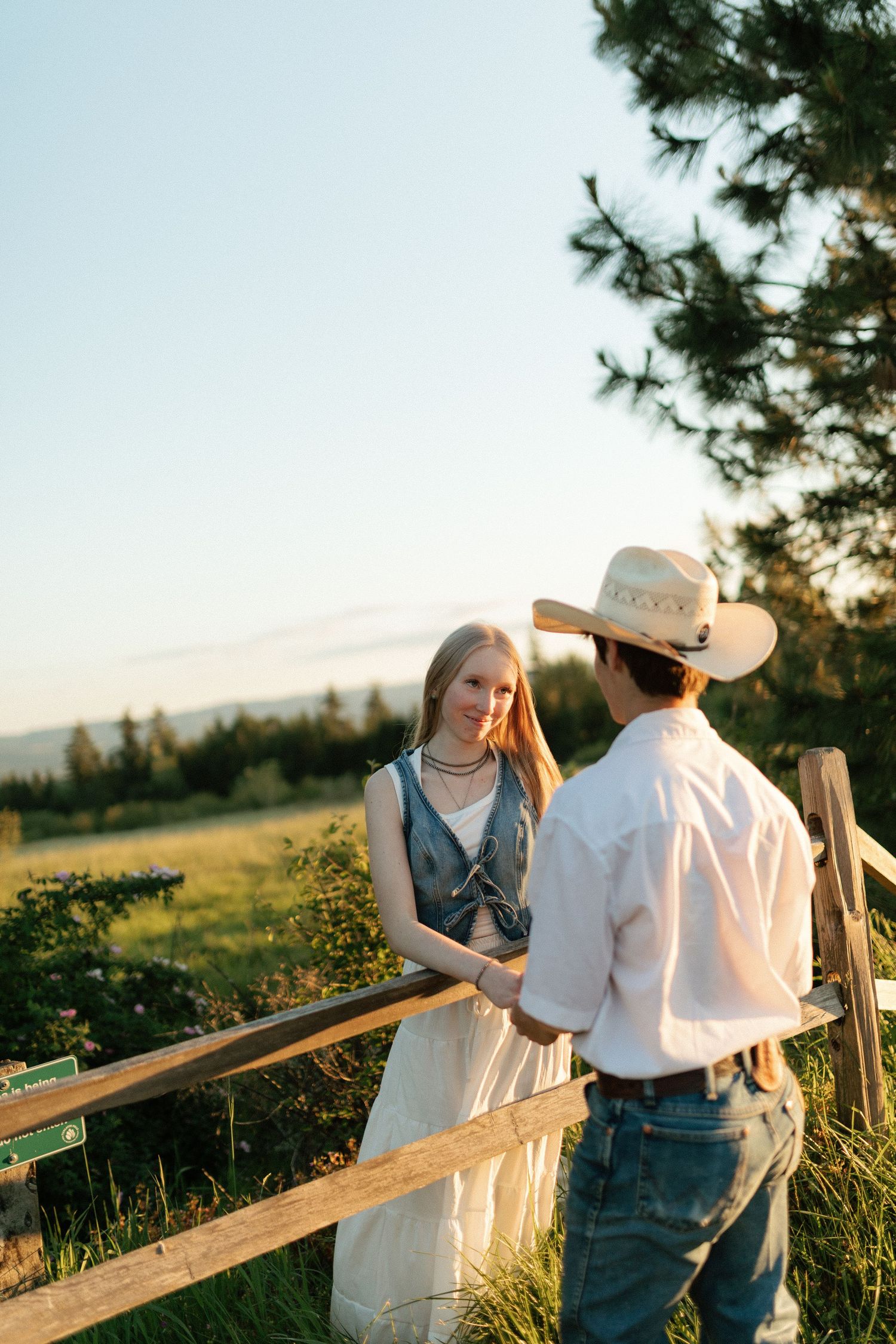 Young couple talking by wooden fence at sunset, woman in white dress, man in cowboy hat and jeans.