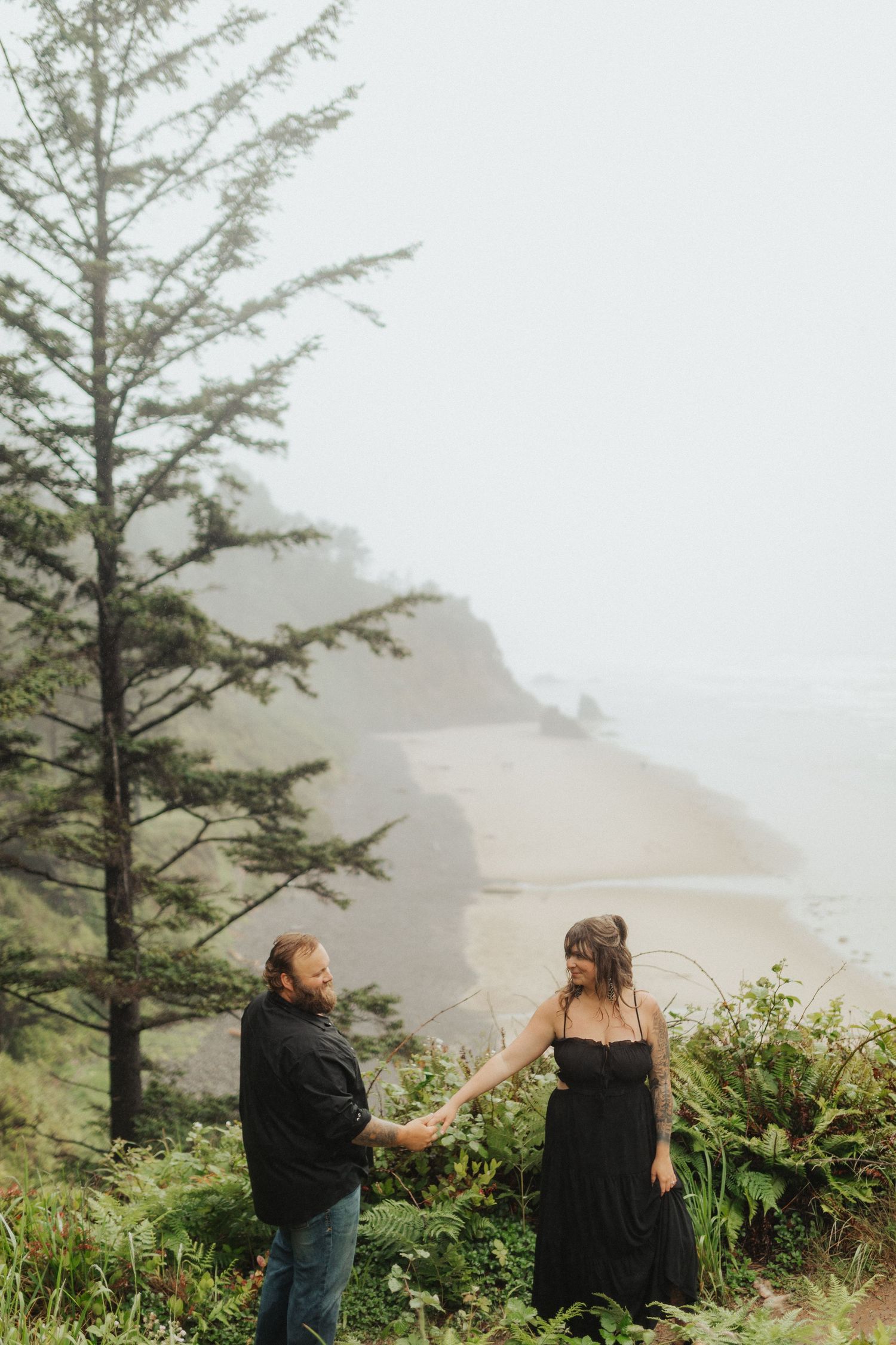Couple holding hands in green ferns on foggy Pacific Northwest coastal overlook with beach below.