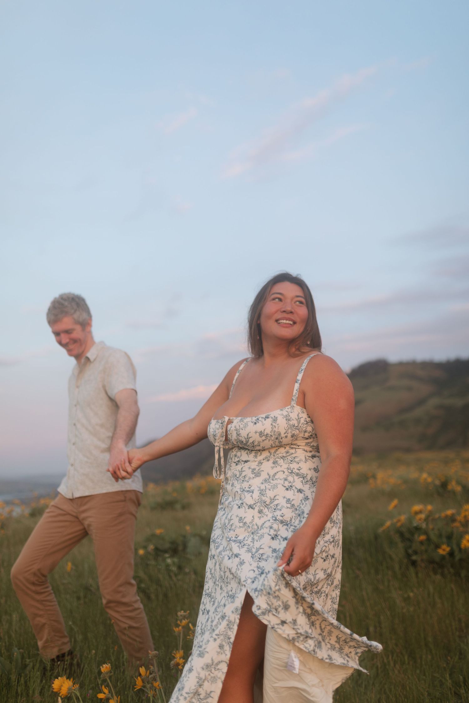 Joyful woman in floral dress leading her partner by hand through a field of yellow wildflowers at dusk.