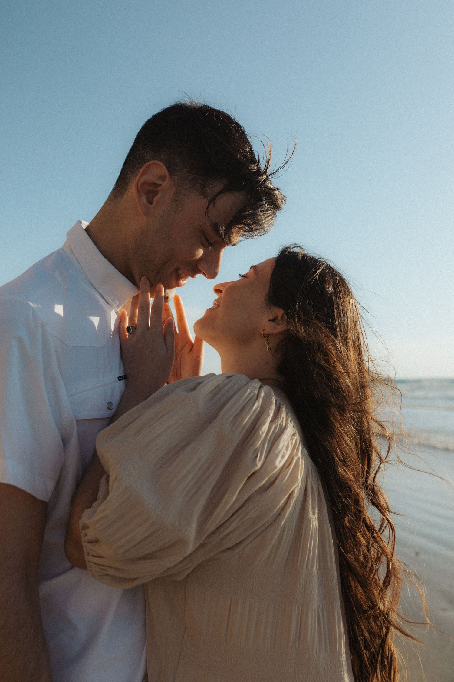 Romantic couple almost kissing on beach at sunset, woman in beige dress and man in white shirt holding hands.