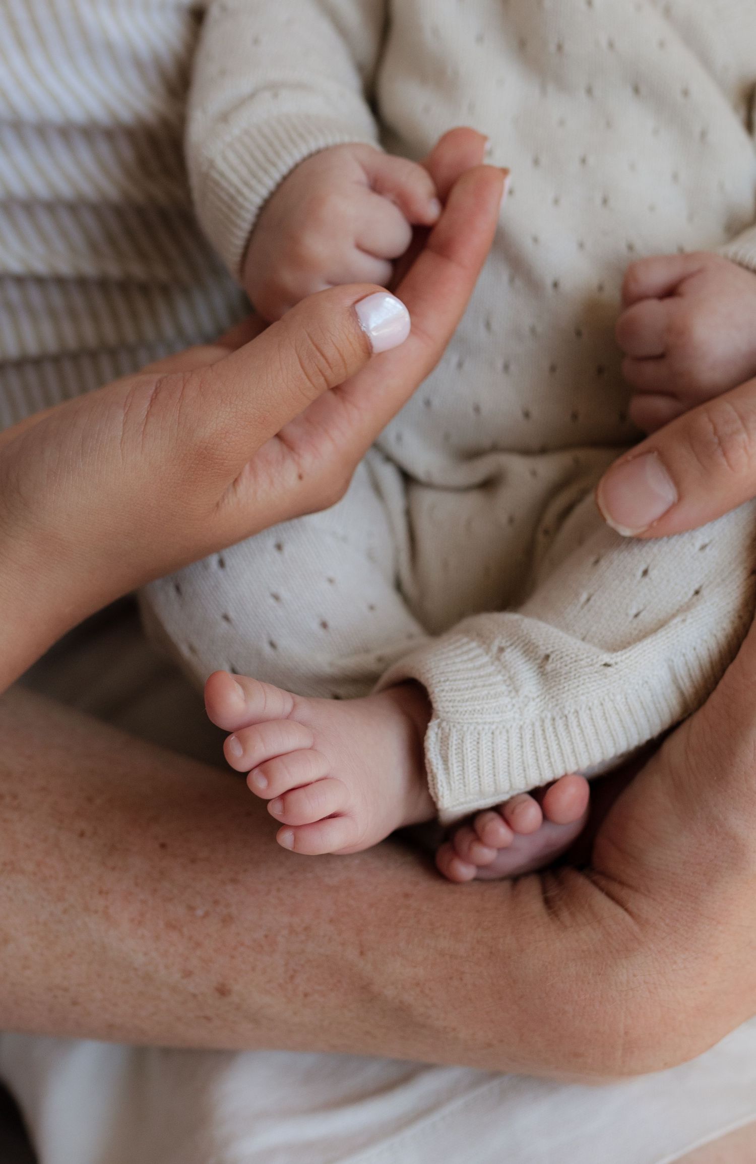 Close-up of newborn baby feet and tiny hands cradled in parent's arms wearing beige dotted onesie.