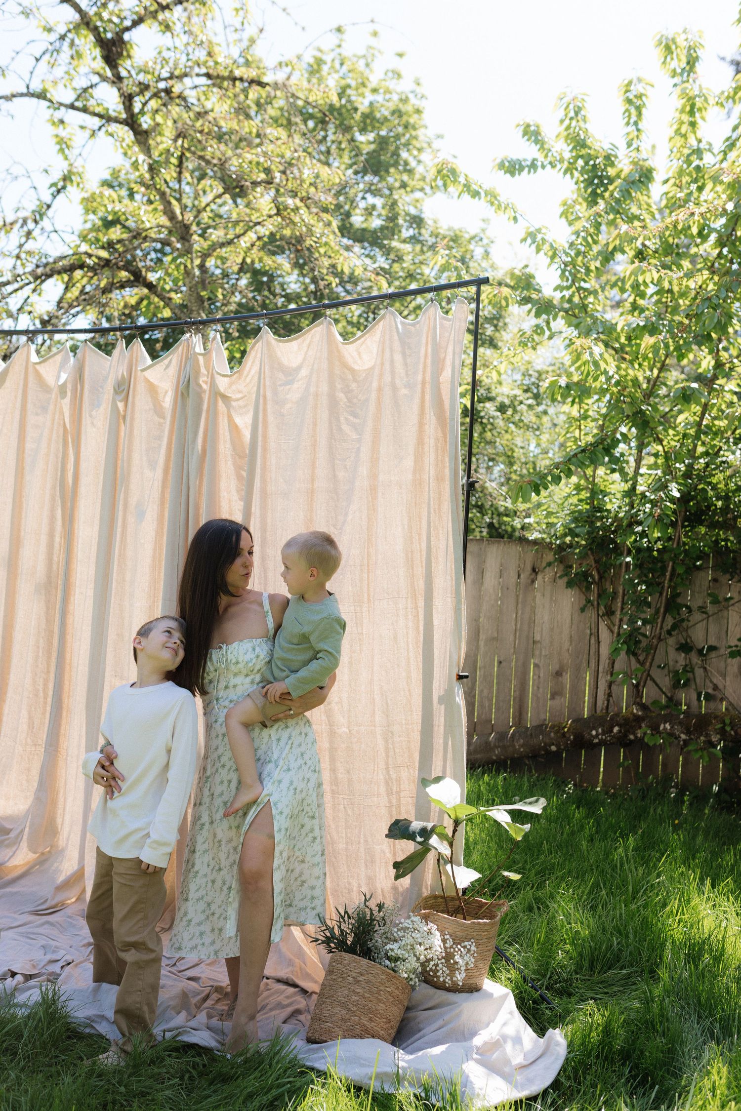A mother holds her toddler while standing with her son in a sunlit garden with a draped backdrop.