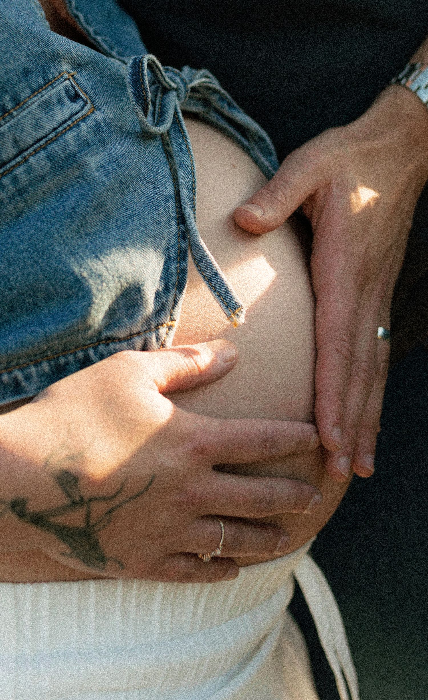 Close-up of hands cradling a pregnant belly, with a denim jacket and anchor tattoo visible.