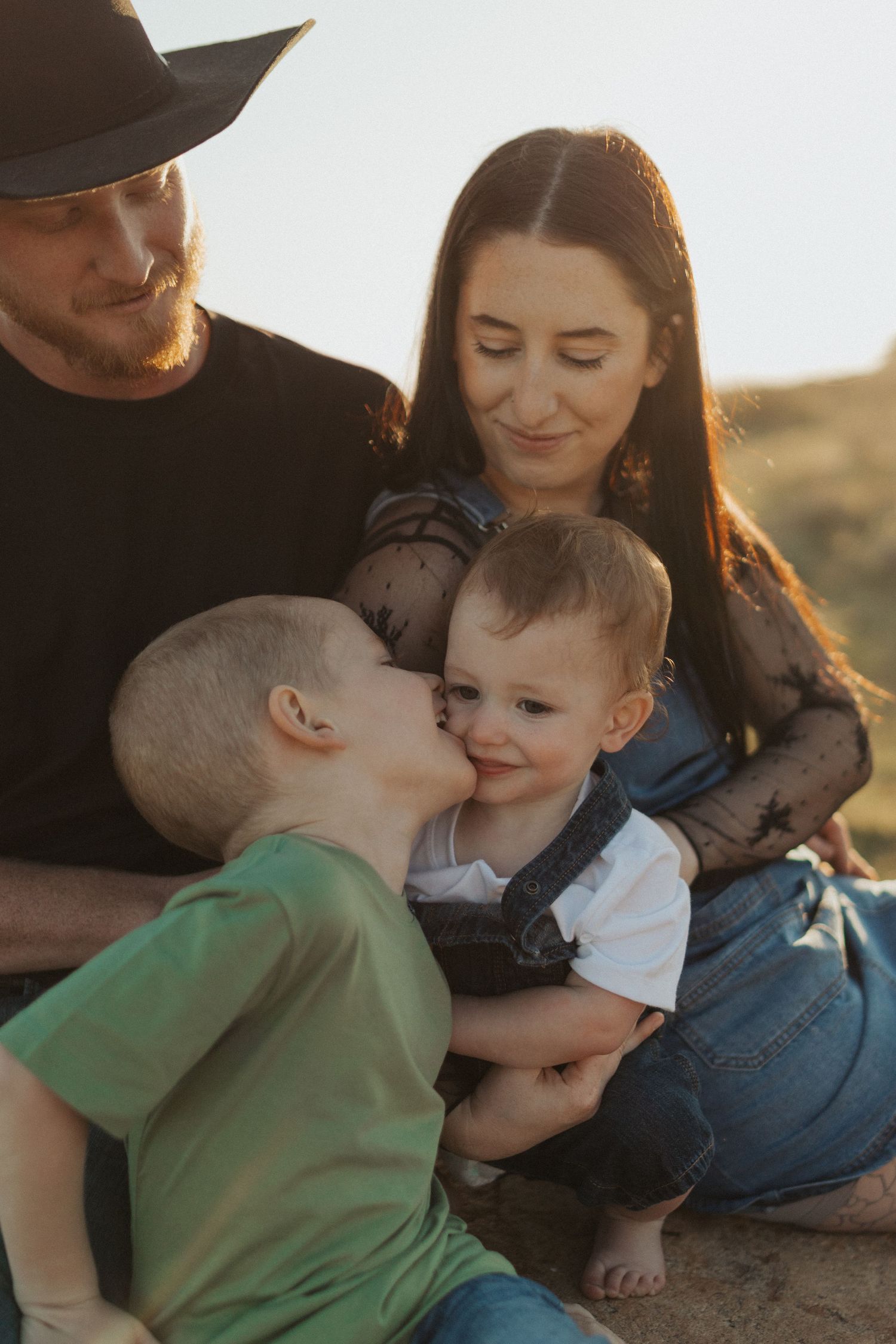 Family portrait with parents and two young boys sharing a sweet kiss outdoors in golden sunlight.