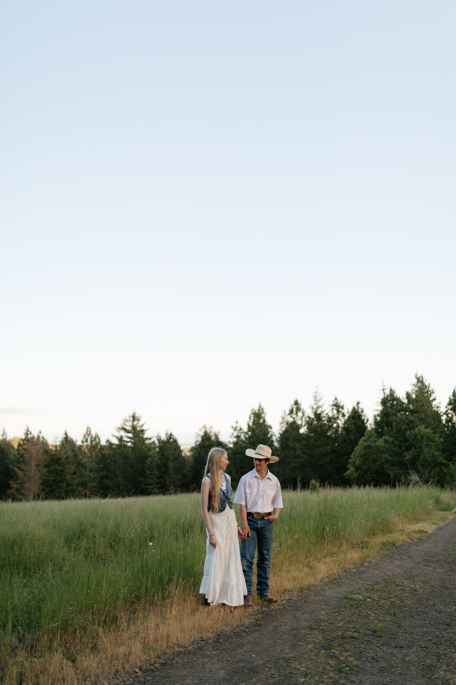 Couple holding hands on a rural path at dusk, woman in white dress, man in cowboy hat.