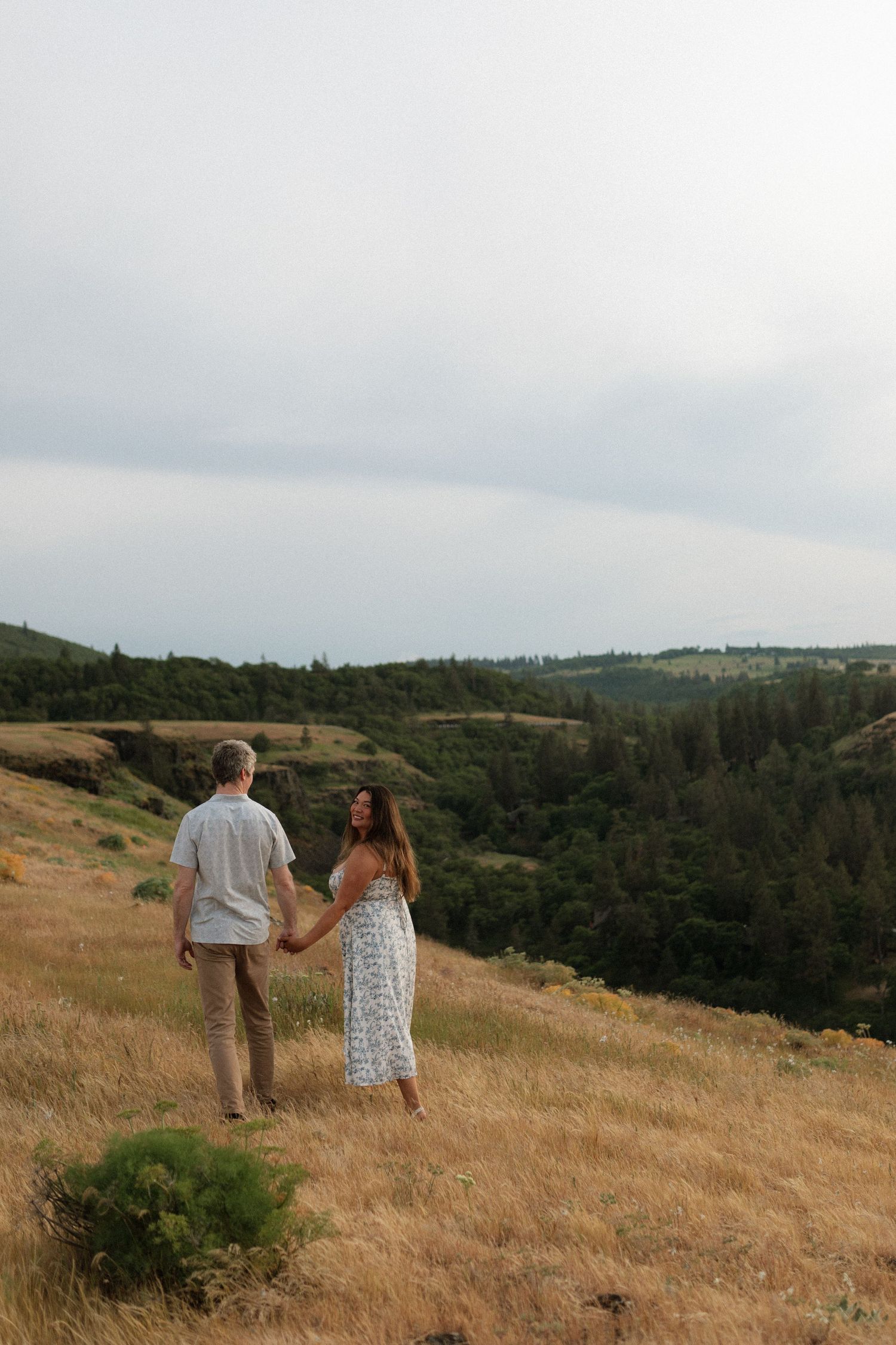 Couple holds hands walking through golden hillside meadow with scenic mountain valley backdrop.