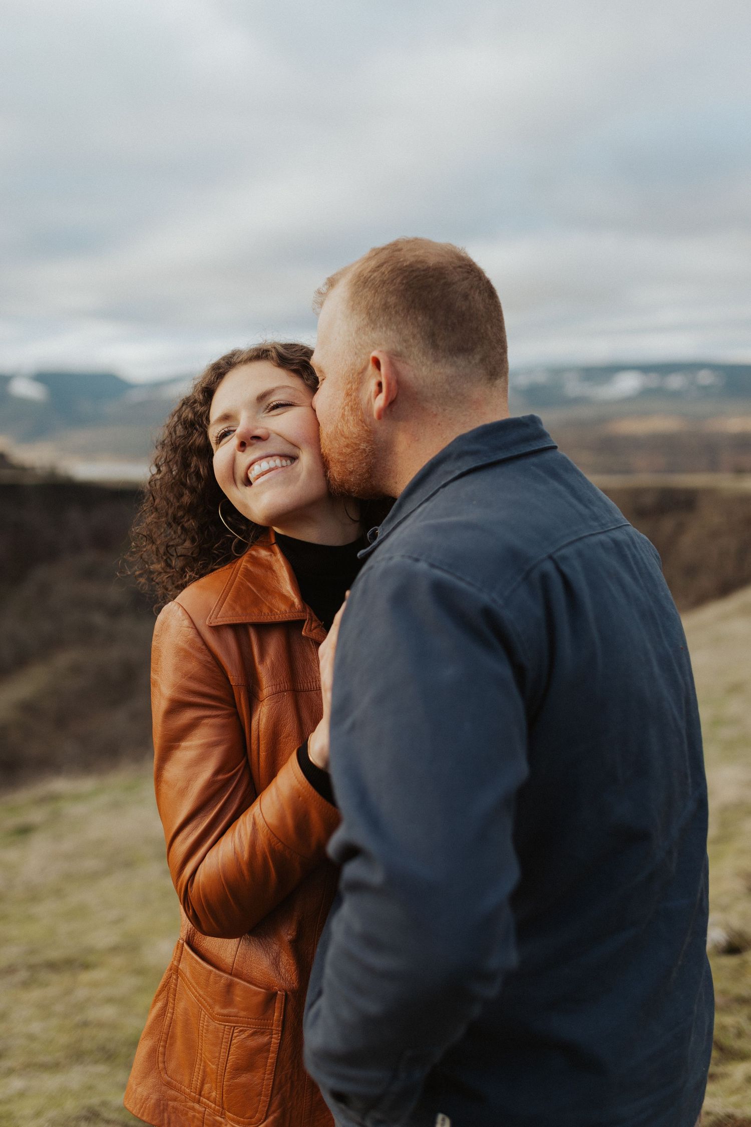 Smiling woman in brown leather jacket receives a kiss on the cheek from her partner with mountain views behind them.