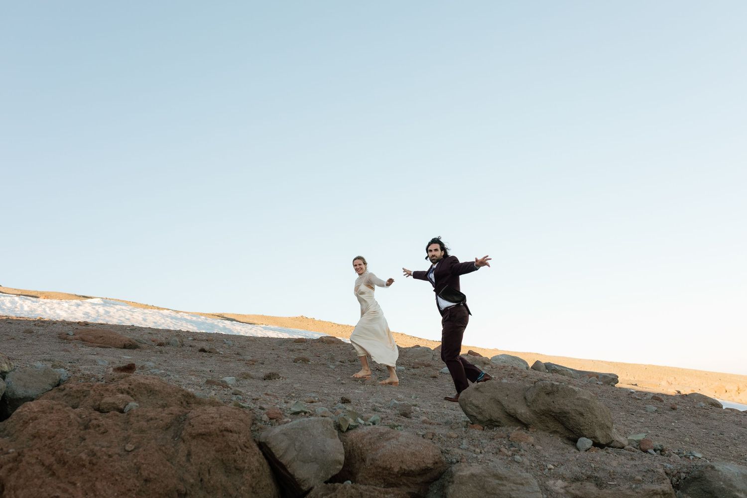 Joyful bride and groom leap playfully on rocky terrain beneath a clear blue sky.