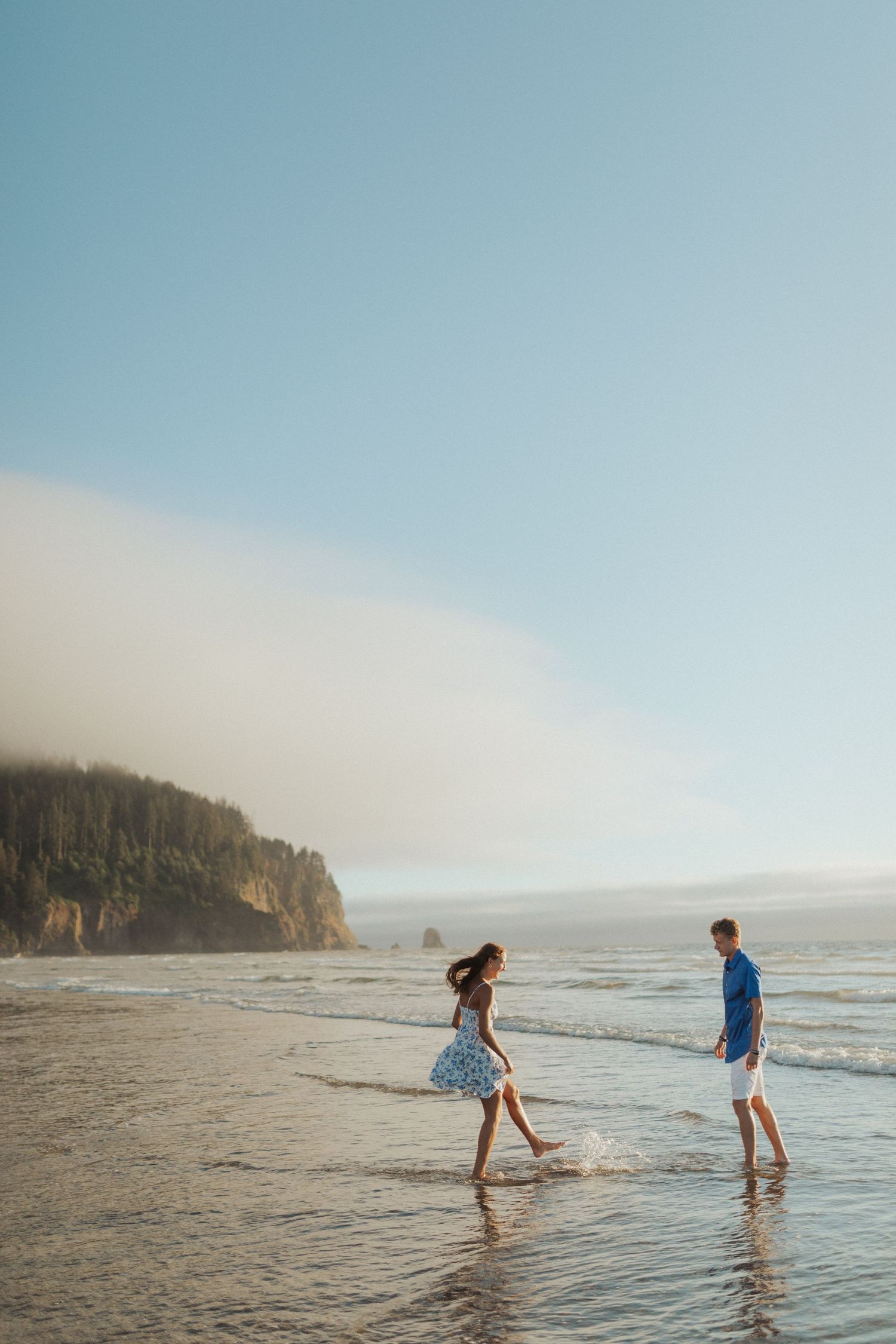 Young couple walking barefoot in shallow ocean water on a misty beach with rocky cliffs.