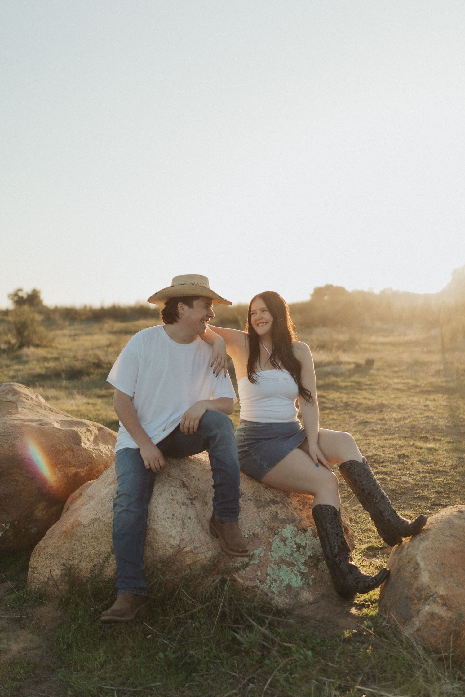Couple sitting on large rocks in a sunny field, smiling at each other in casual western attire.