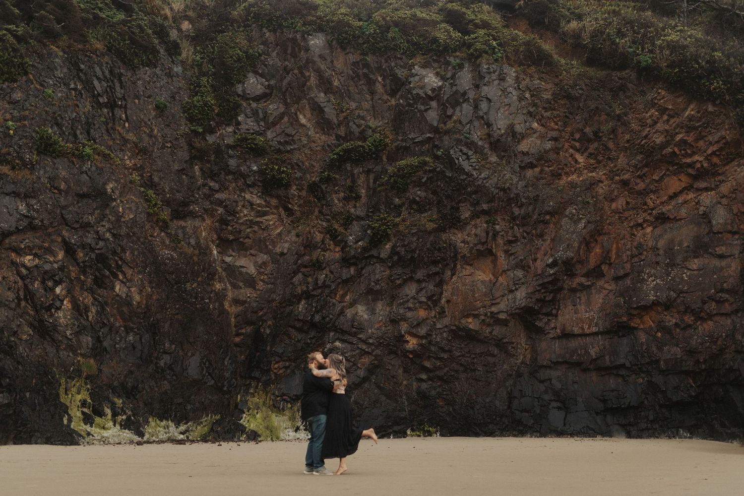 Couple embracing on a wide sandy beach with dramatic rocky cliffs and green foliage behind them.