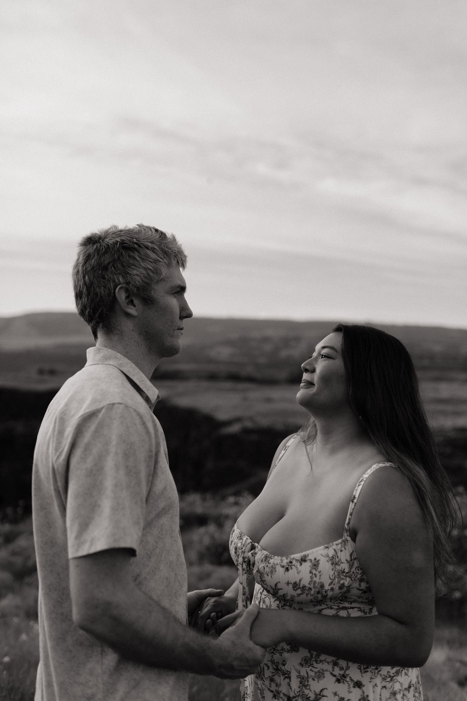 Black and white portrait of couple holding hands and gazing at each other on open hillside.