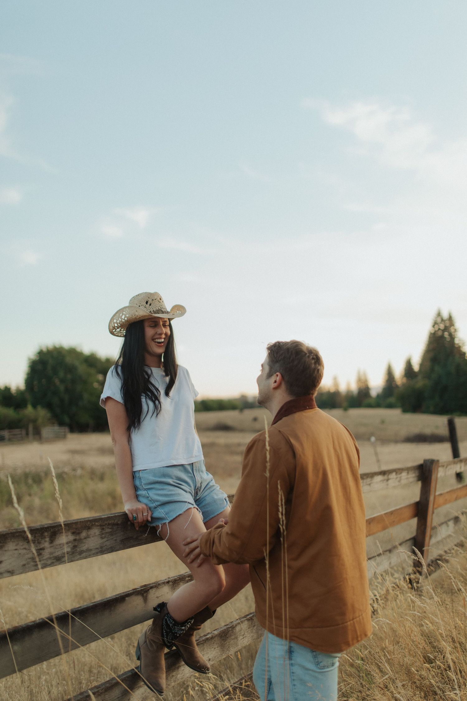 A smiling woman in a cowboy hat sits on a fence holding hands with her partner.