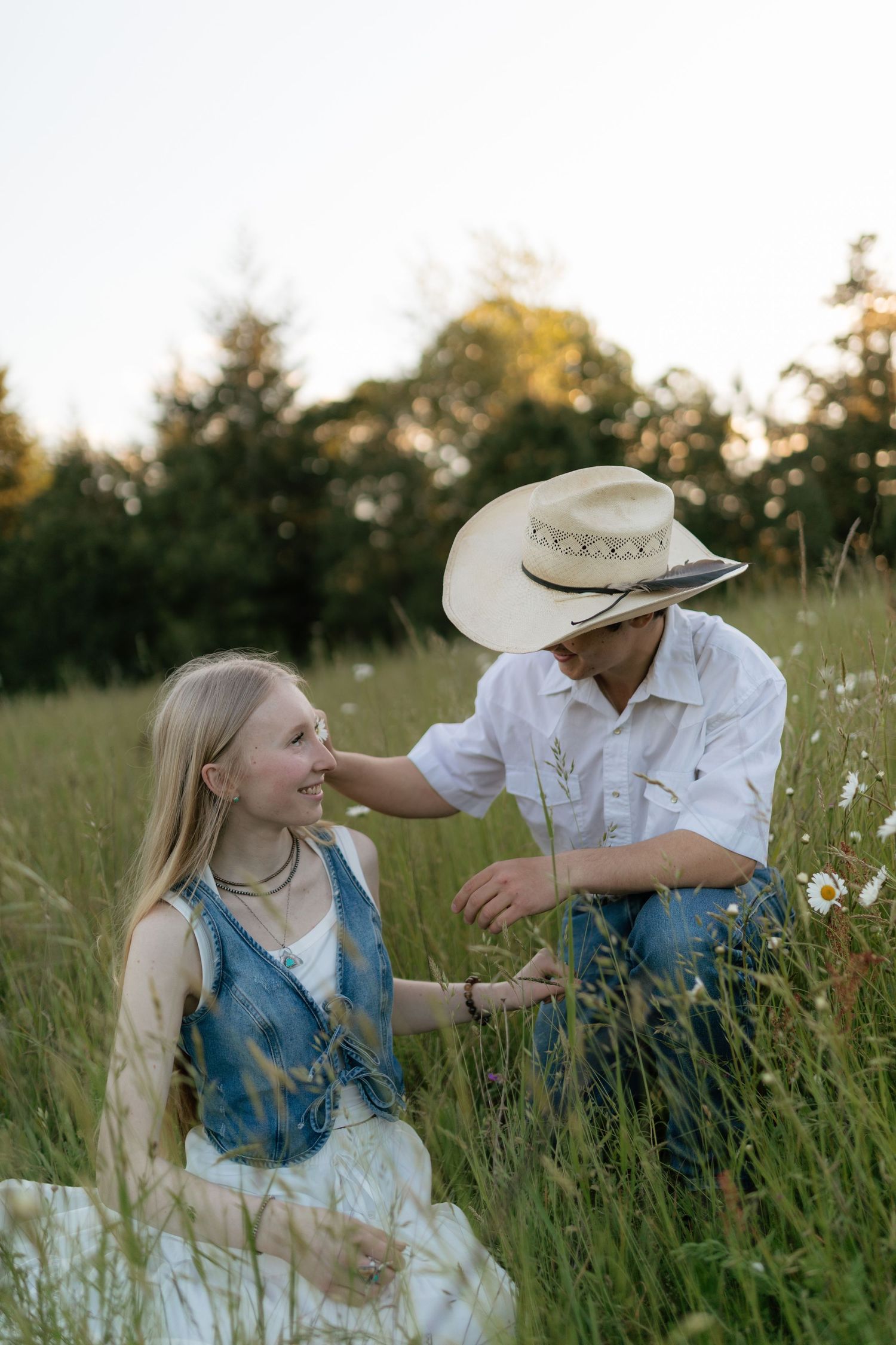 Couple sitting together in tall grass meadow with daisies, man in cowboy hat touching womans hair.