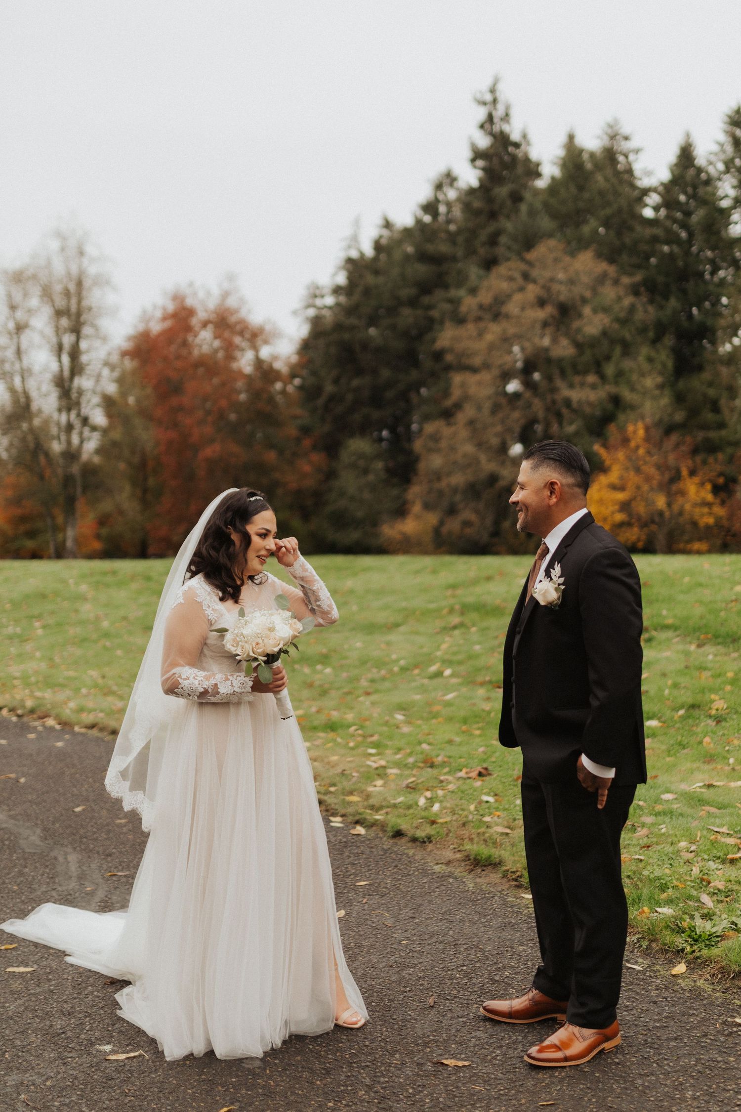 Emotional bride in lace gown with veil faces groom in black suit during first look in autumn park setting.
