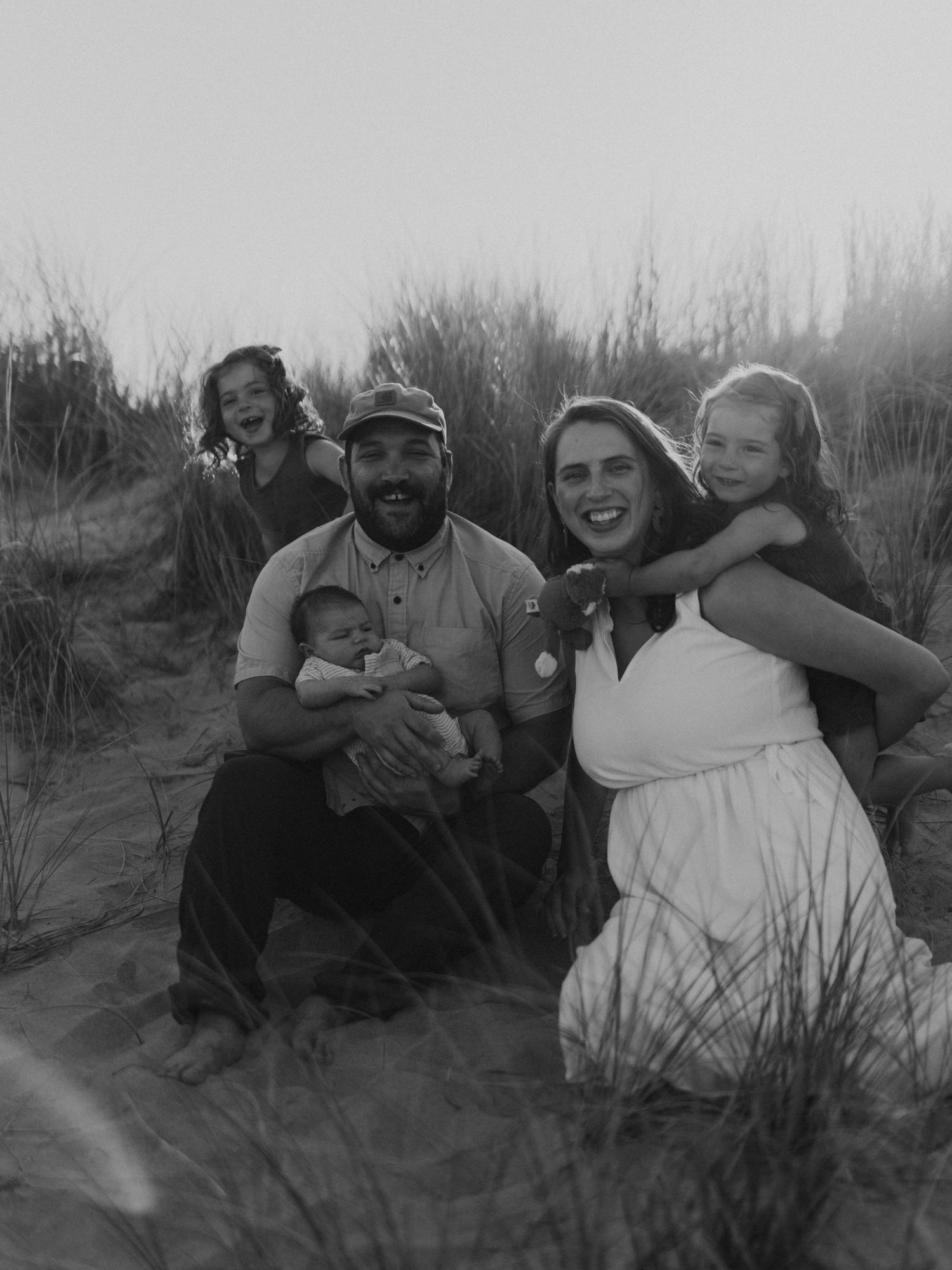 Black and white family photo with parents, newborn, and two daughters sitting in sandy beach dunes.