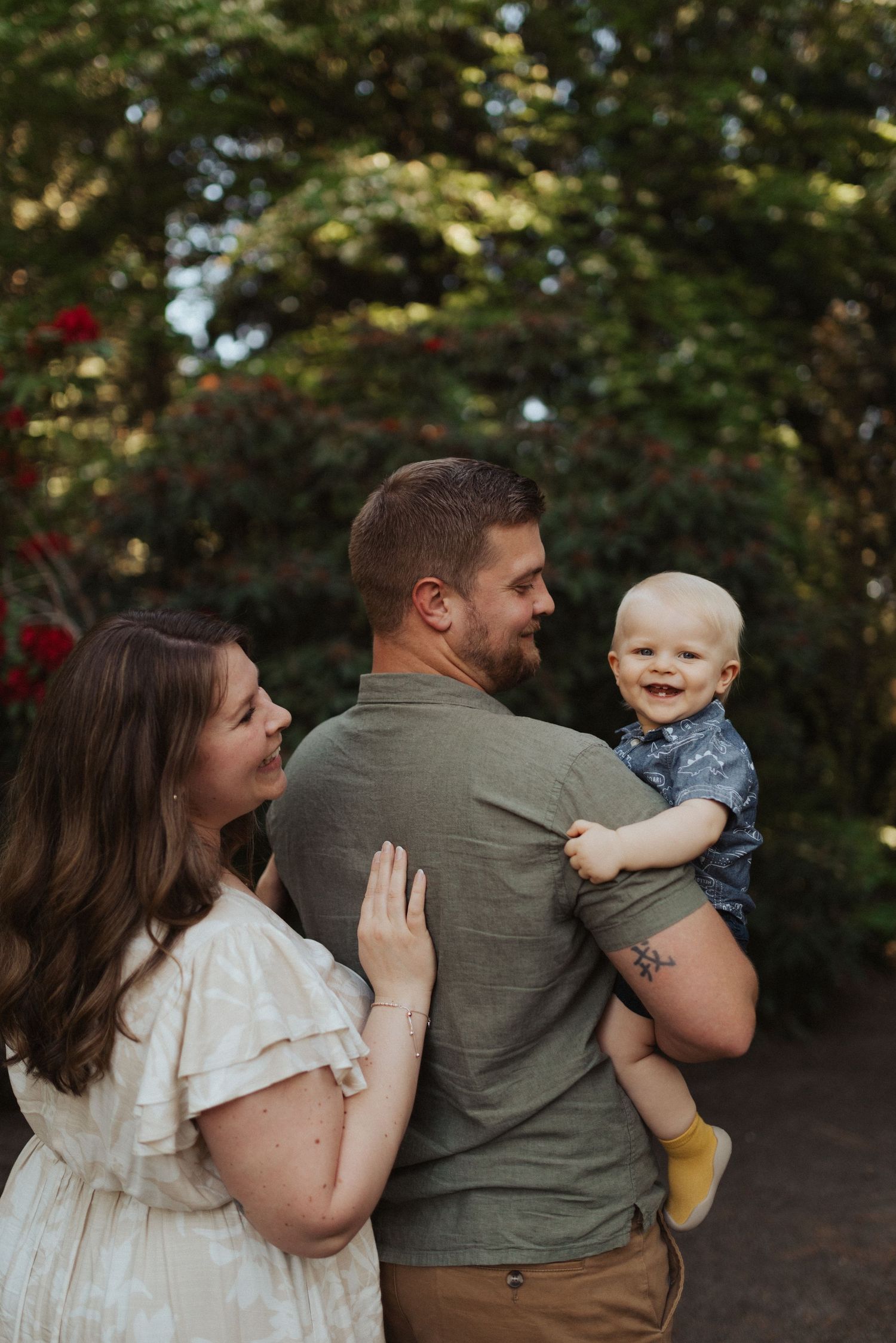 Young couple with smiling baby boy posing outdoors near blooming red rose bushes in garden.