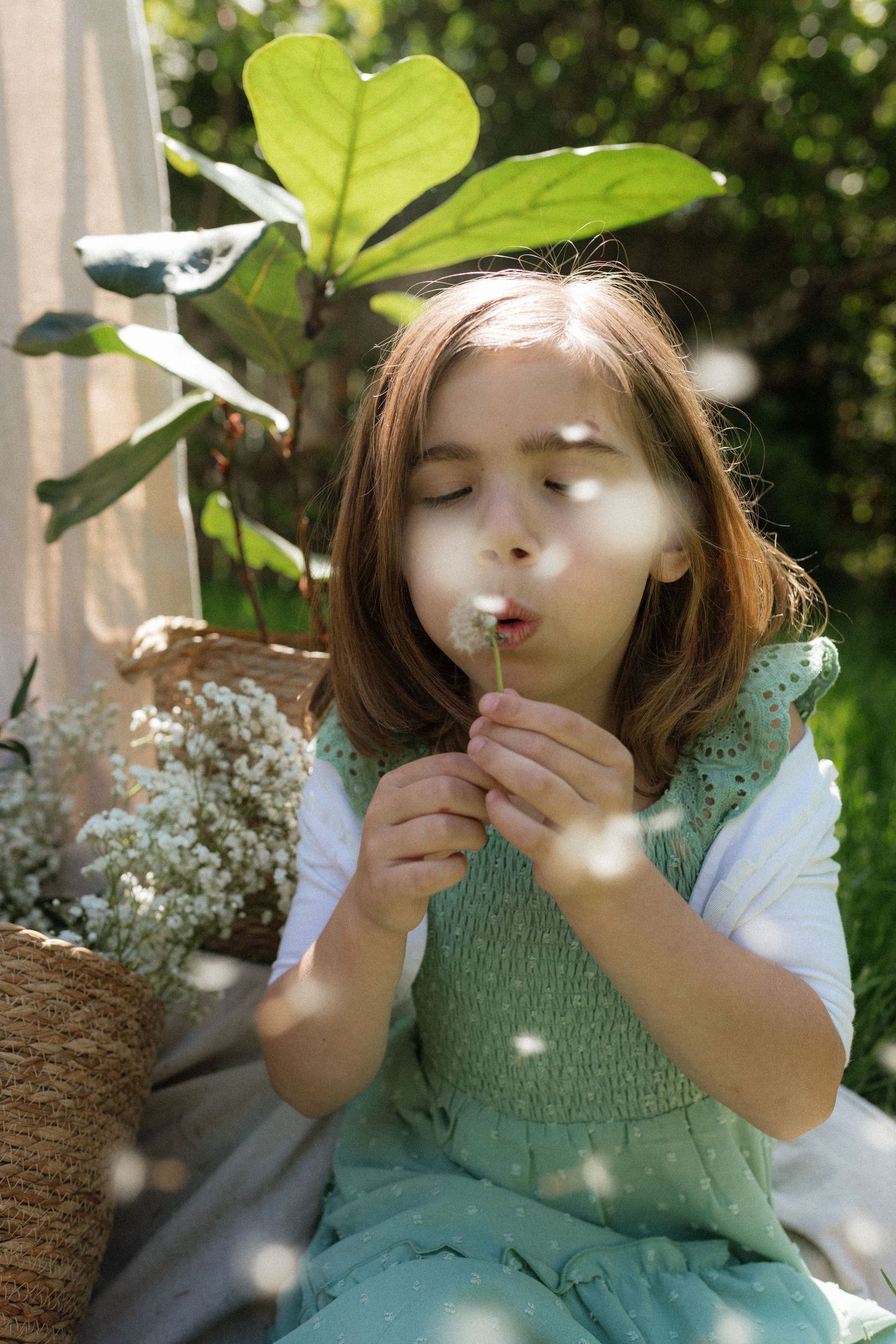 A young girl with brown hair blows a dandelion in a lush garden, wearing a green dress.