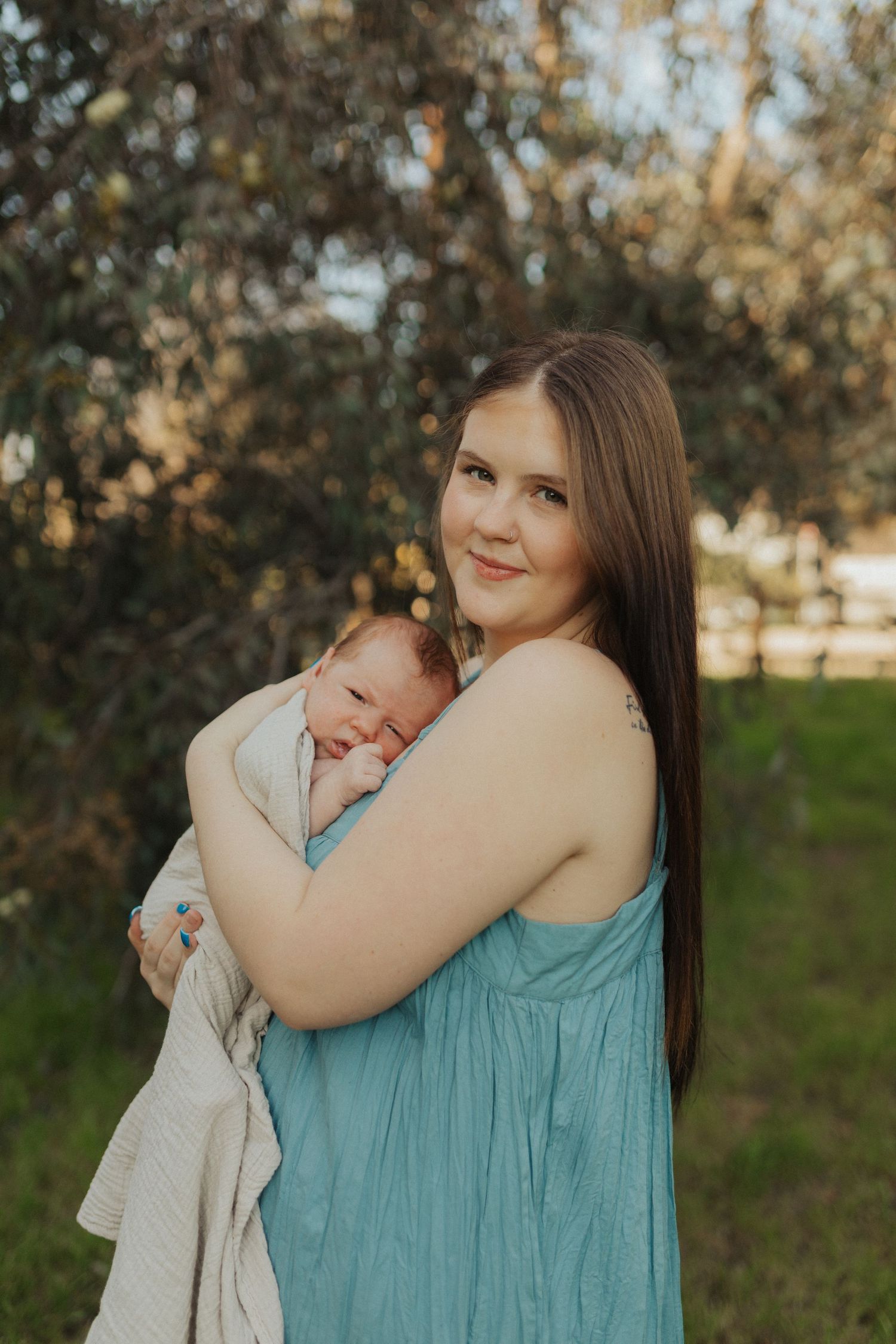 Smiling mother in teal dress holding swaddled newborn baby outdoors in warm golden hour light.