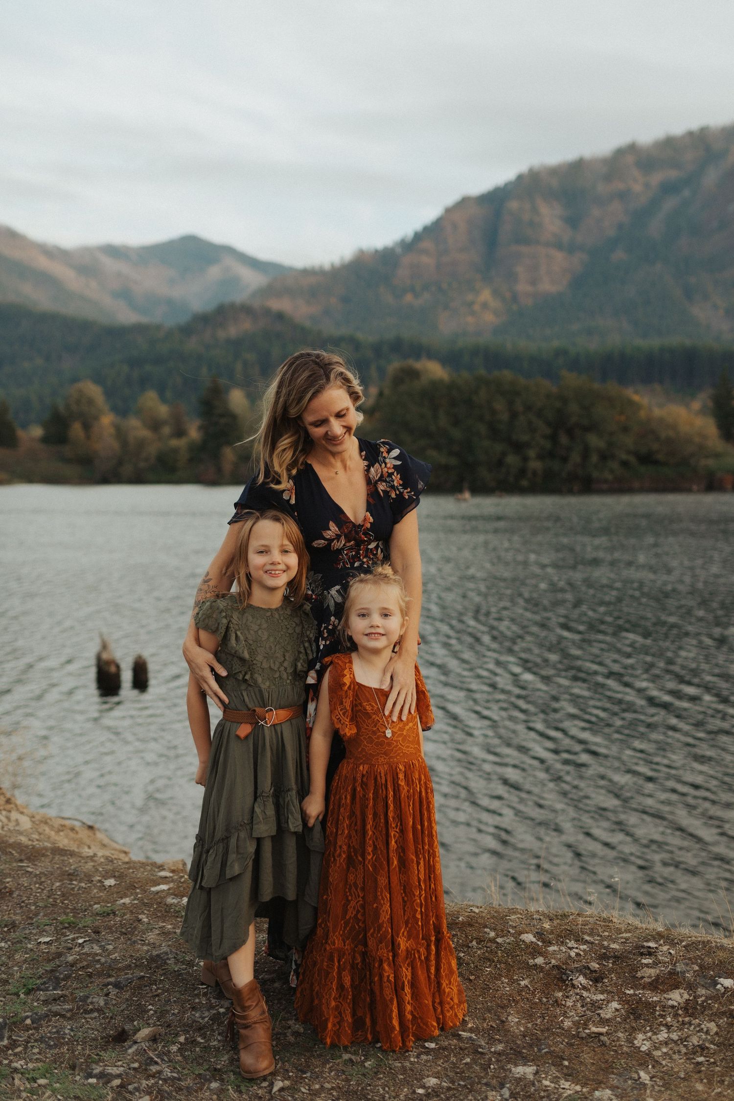 A smiling mother poses with her two daughters in boho dresses beside a serene mountain lake.