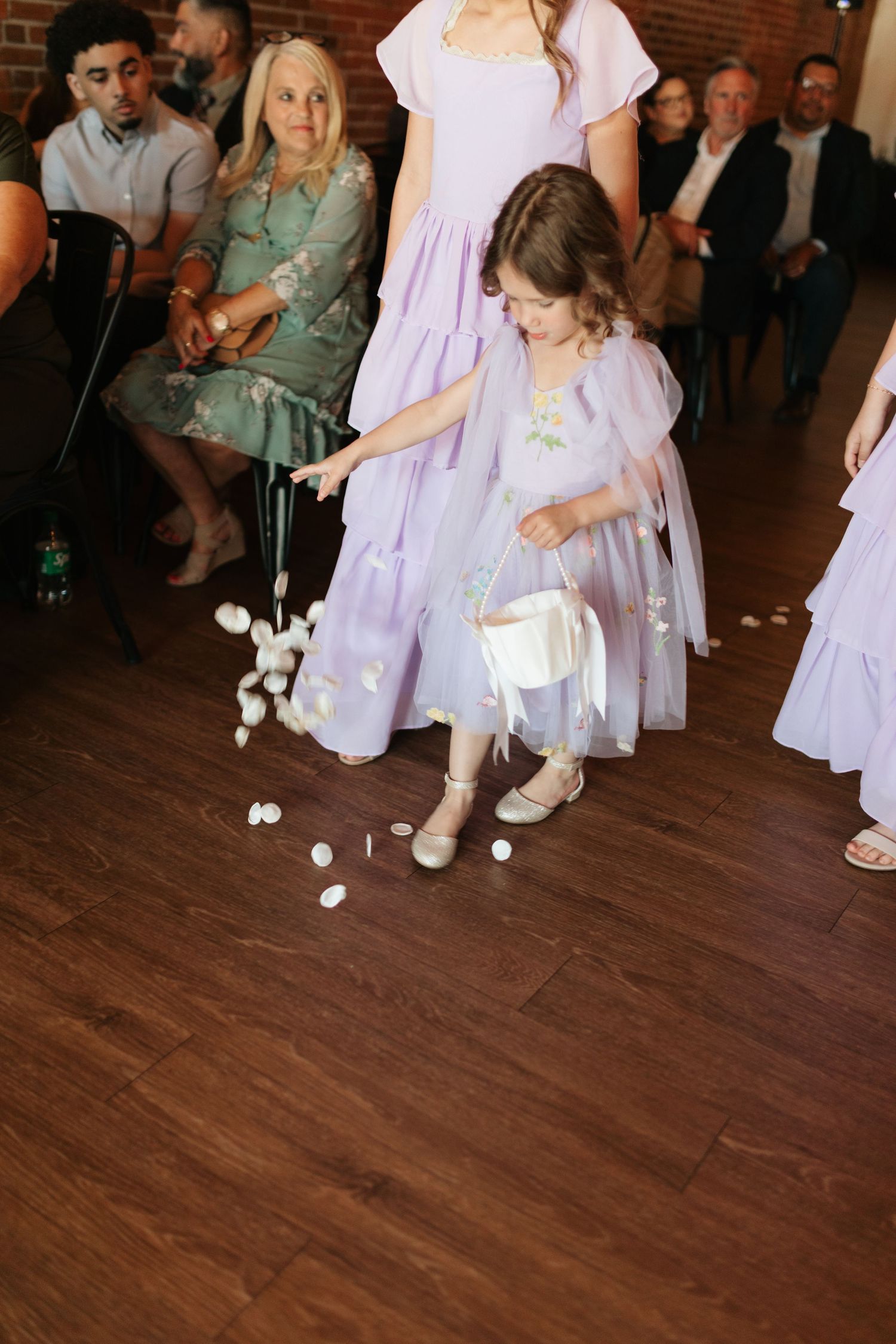 Young flower girl in lavender dress scattering white petals on wooden floor during wedding ceremony.