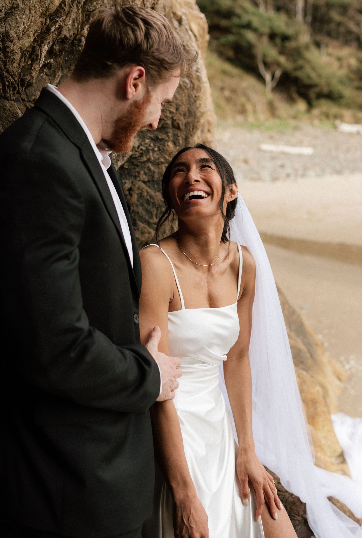 Bride laughs joyfully while groom gazes at her lovingly, standing against rocky coastal backdrop on beach.
