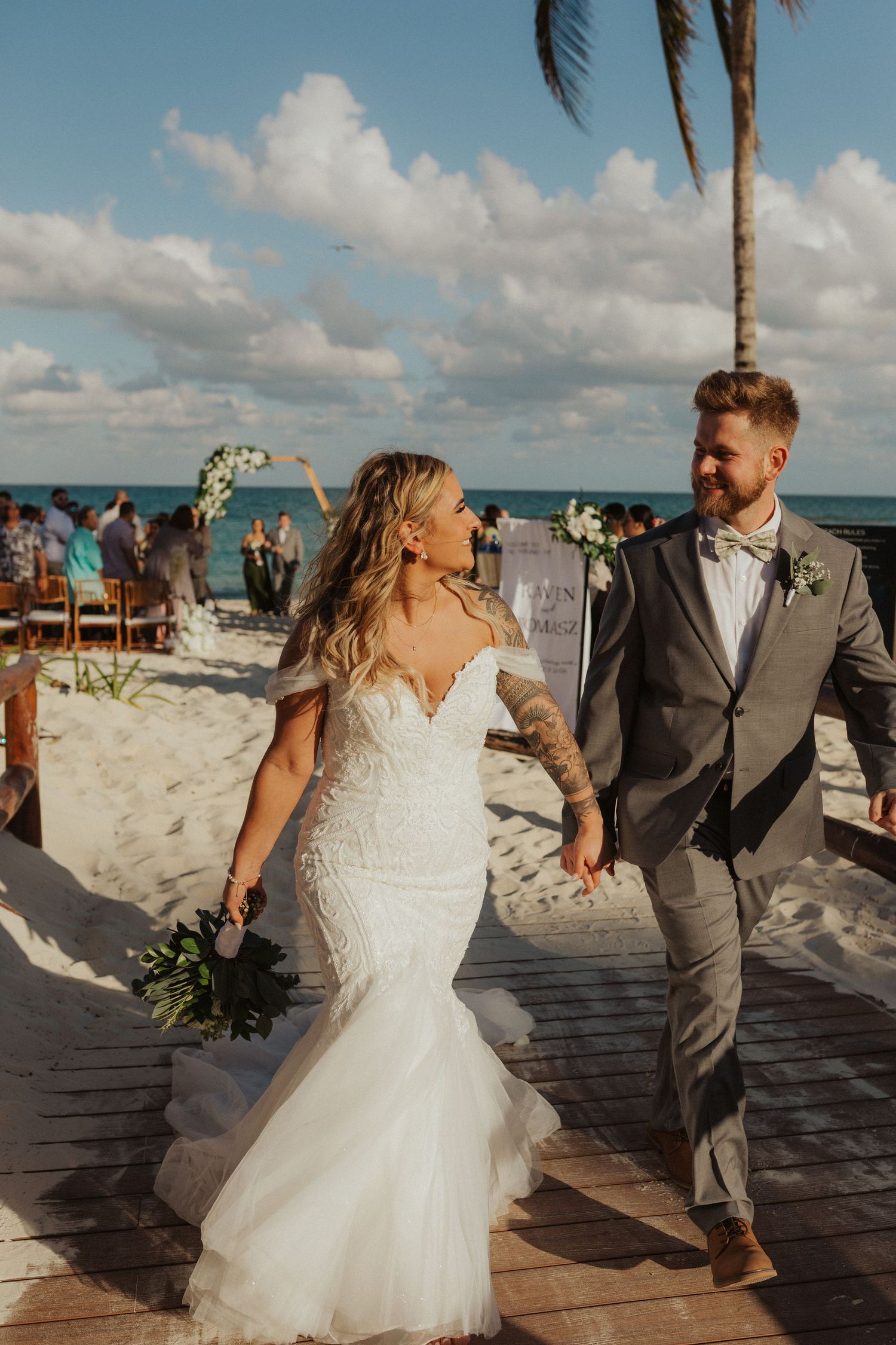 Joyful newlyweds walking down beach boardwalk aisle after ceremony with ocean and palms behind them.