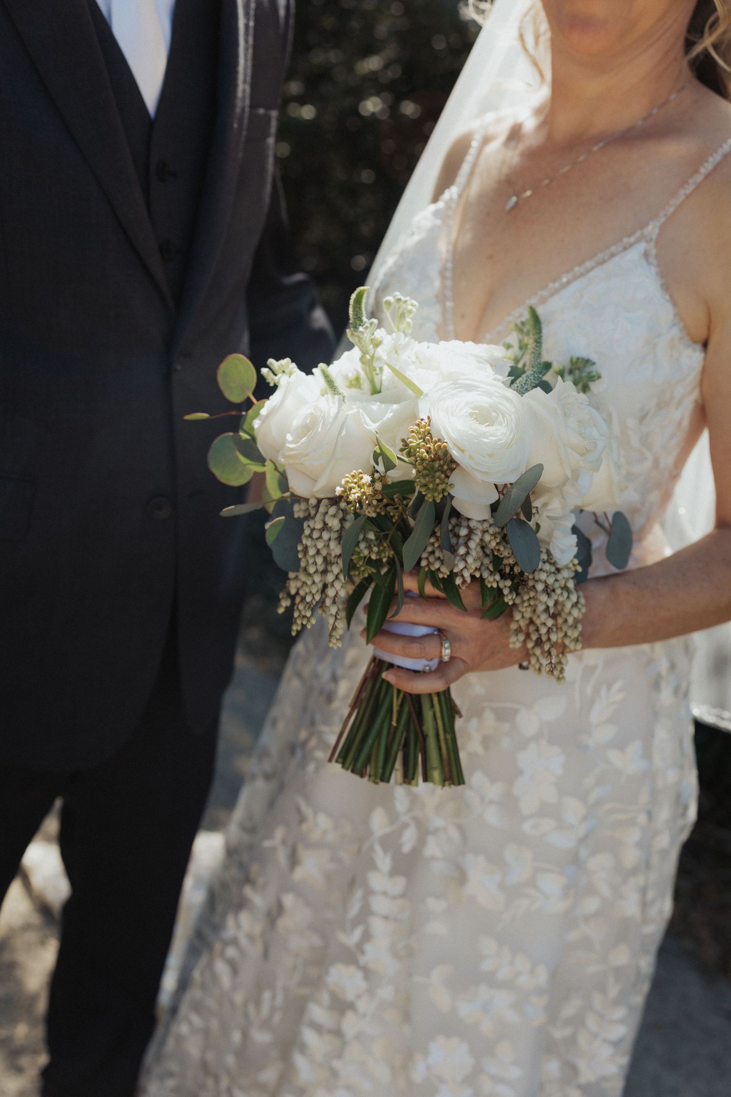 Bride holding a white rose and eucalyptus bouquet with gold accents beside groom in black suit.