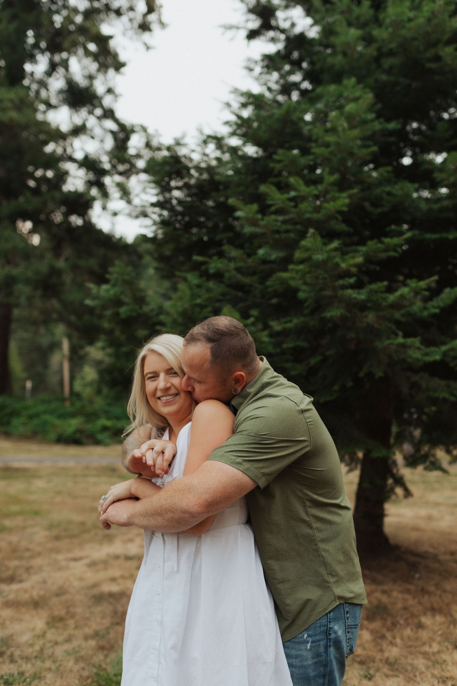 Man in green shirt embracing and kissing woman in white dress outdoors among tall evergreen trees.
