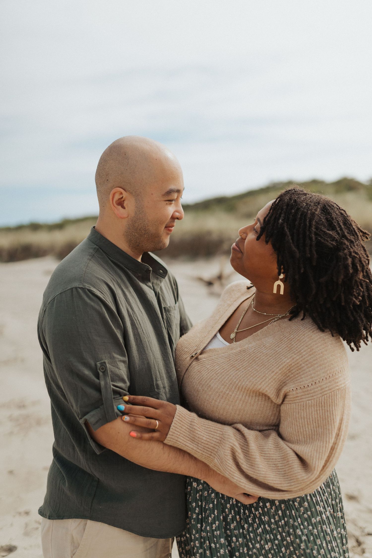 Interracial couple sharing a tender moment together on a sandy beach with dune grass behind them.