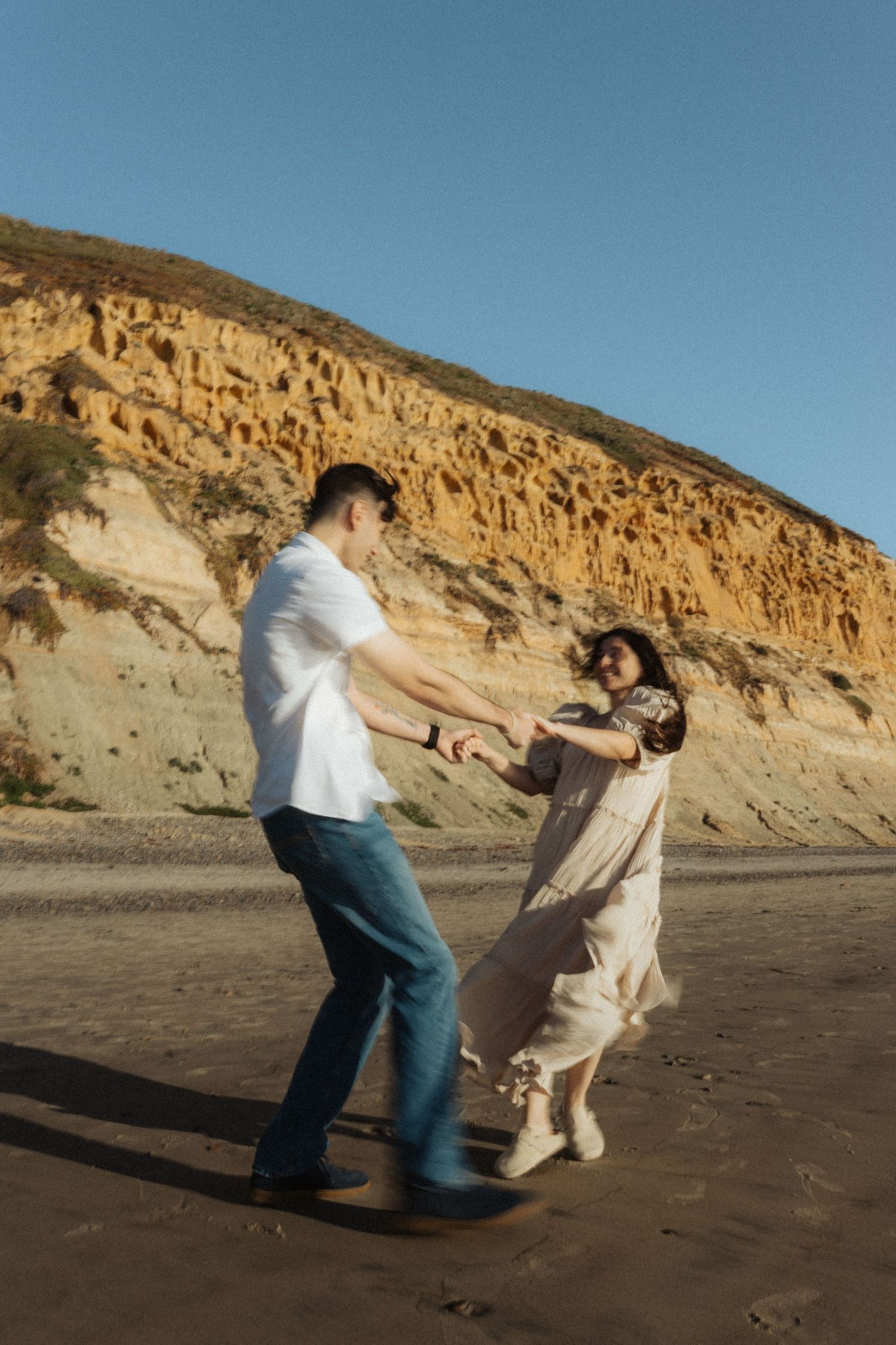 Joyful couple dancing and spinning on sandy beach with dramatic golden cliffs in the background.