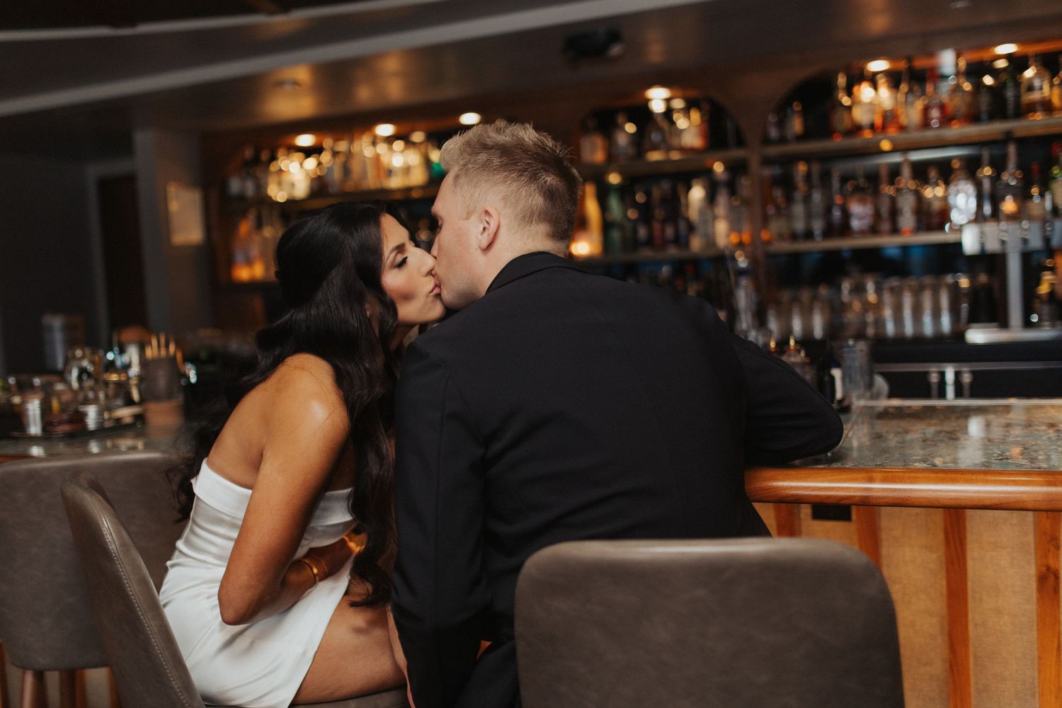 Couple sharing a kiss at an upscale bar, woman in white dress and man in black suit on bar stools.