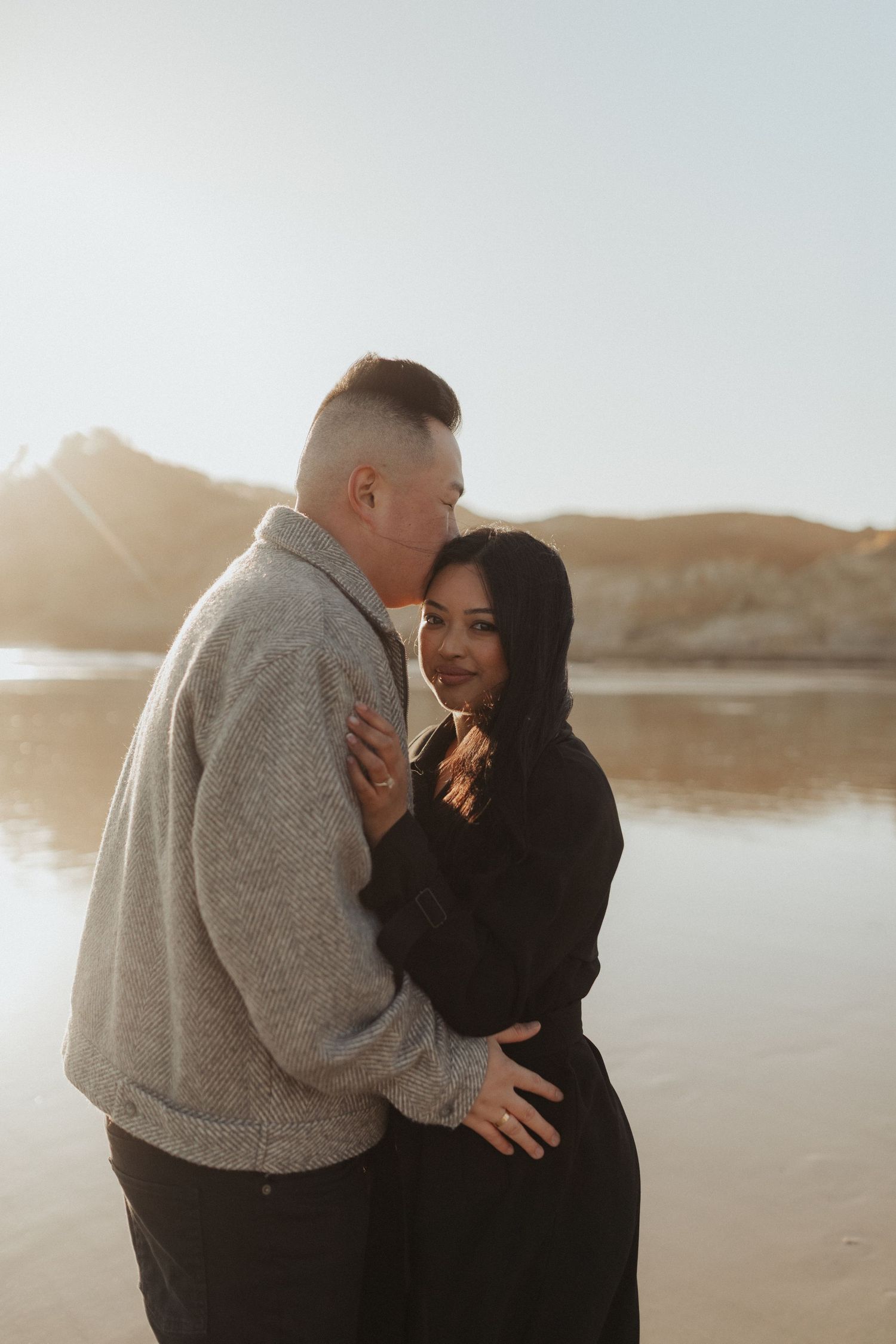 Couple sharing a tender moment on glowing sunset beach, woman smiling while man kisses her cheek.