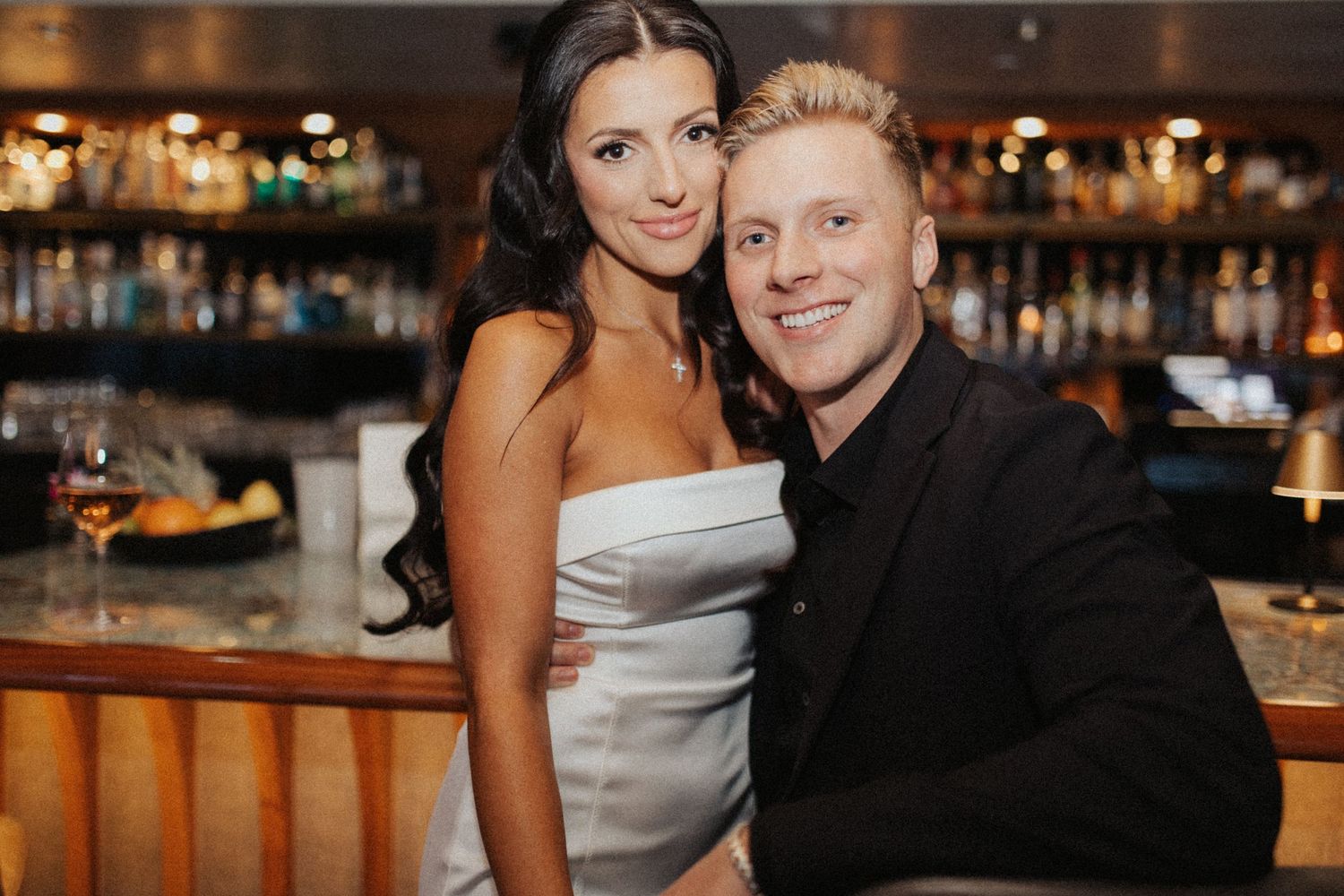 Smiling couple posing together at a stylish bar, woman in silver strapless dress and man in black suit.
