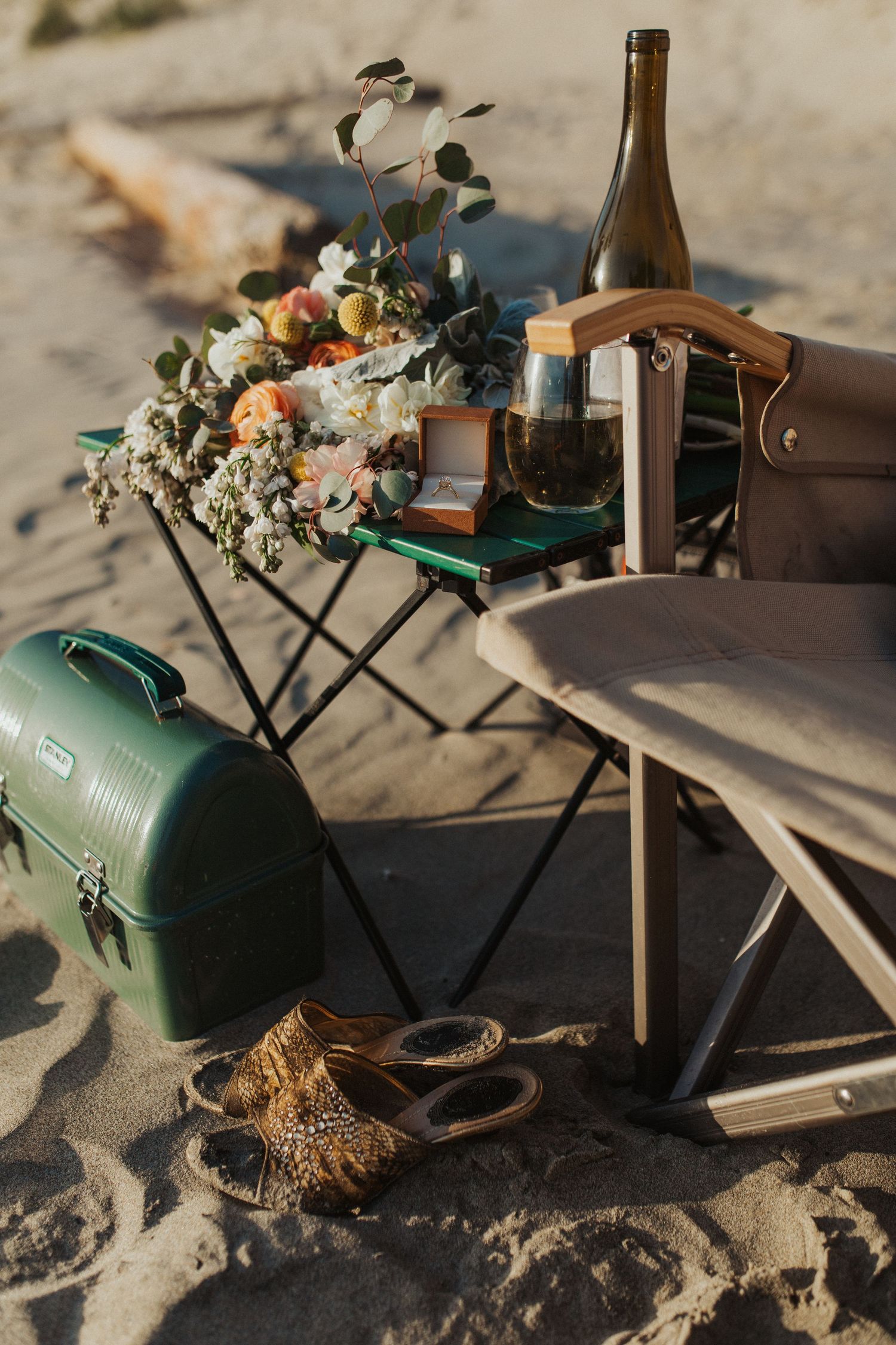 Beach picnic setup with flowers, wine bottle, ring box, and vintage cooler on sandy shore.