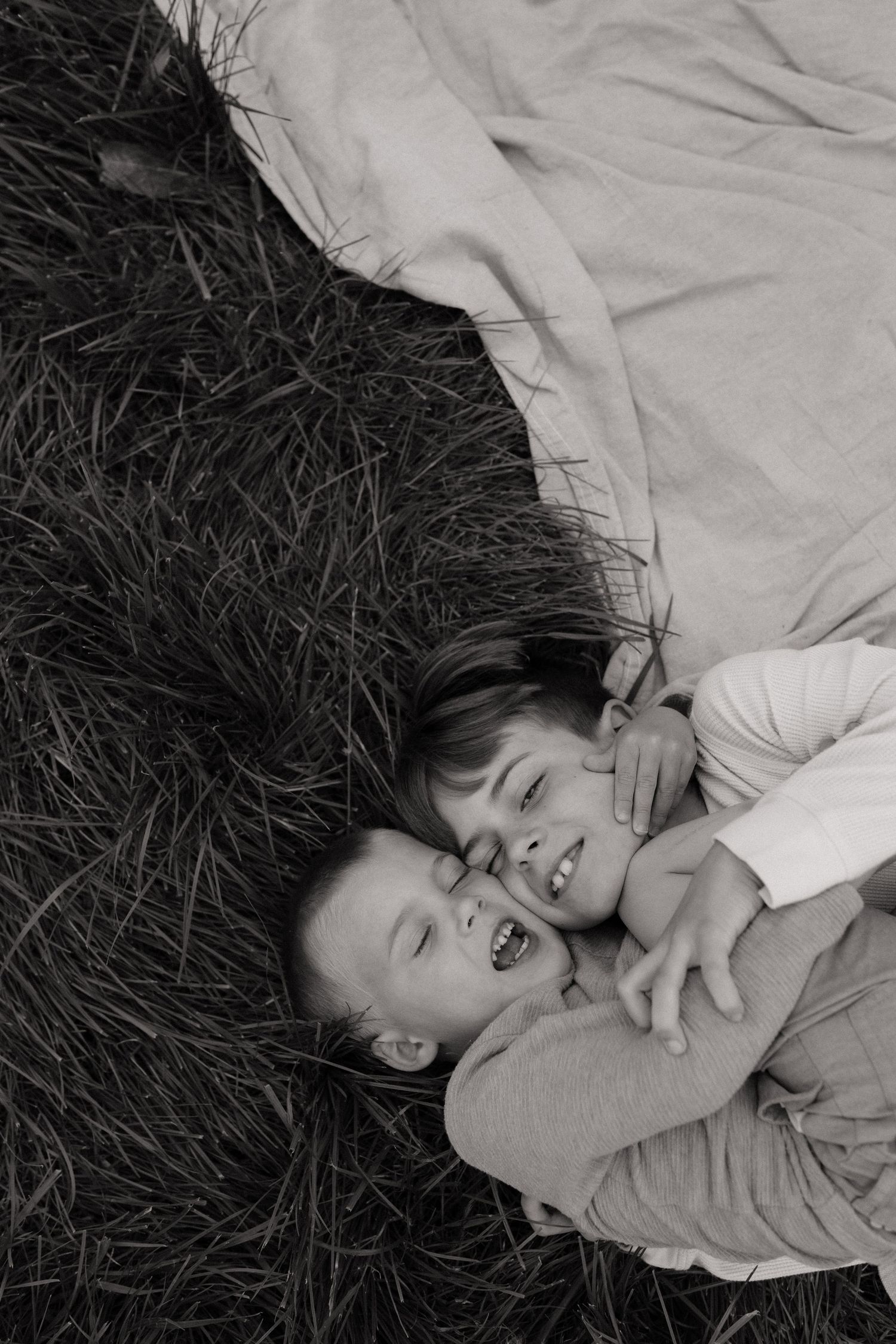 Two laughing children lie together on grass in a charming black and white portrait photo.