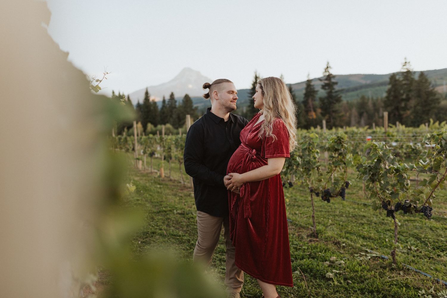 Couple sharing a tender moment in a lush vineyard with mountain views during a maternity photo session.