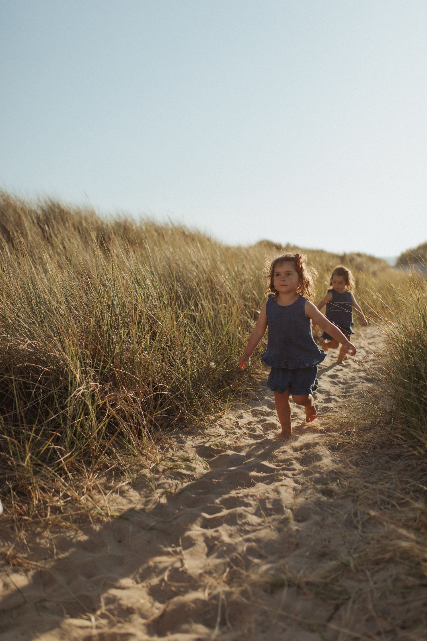 Two young girls in matching blue dresses walking barefoot along a sandy beach dune path.