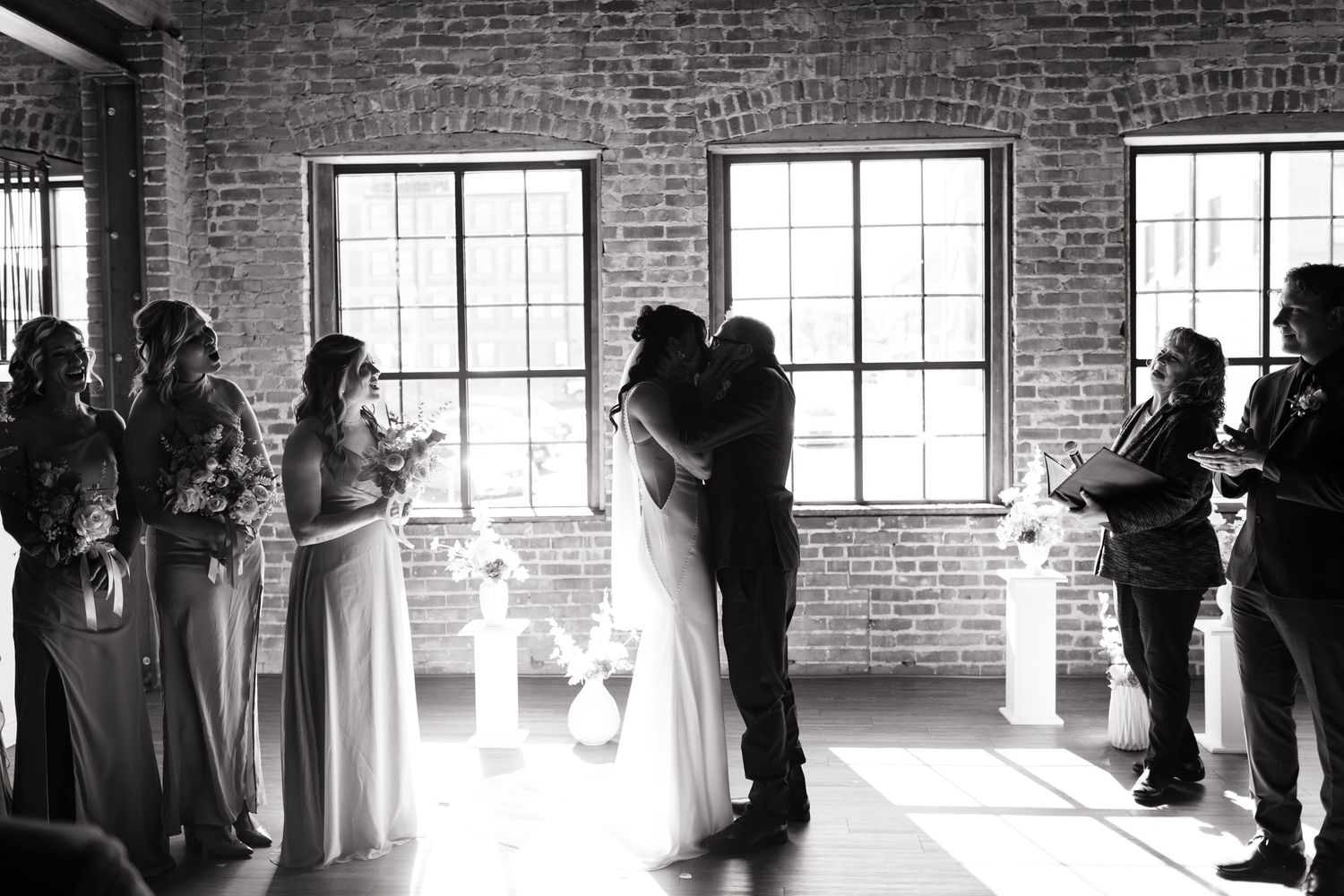 Black and white photo of bride and groom sharing first kiss at altar with wedding party.